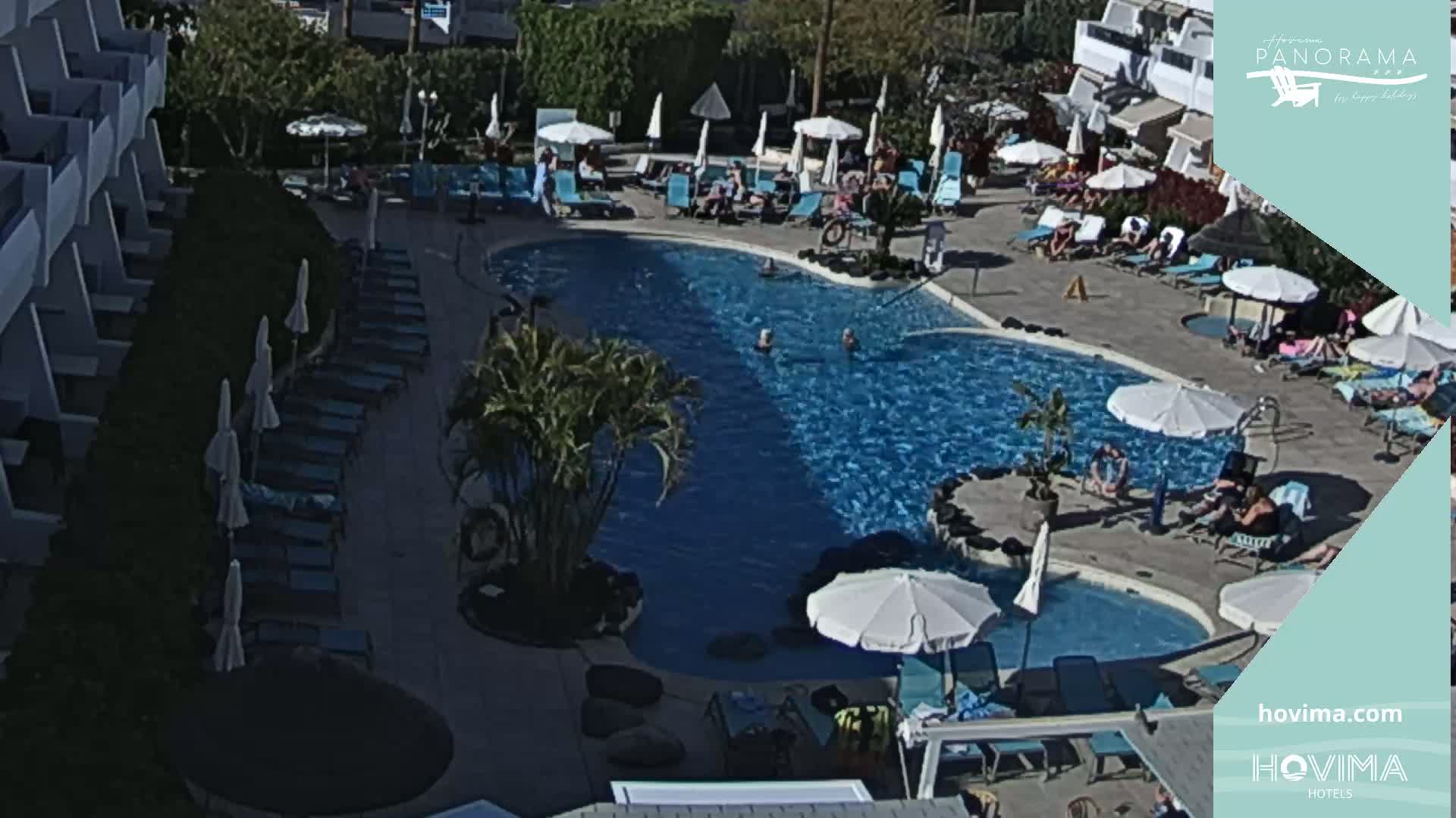 An outdoor swimming pool surrounded by lounge chairs and umbrellas, with people swimming and sunbathing on a sunny day.
