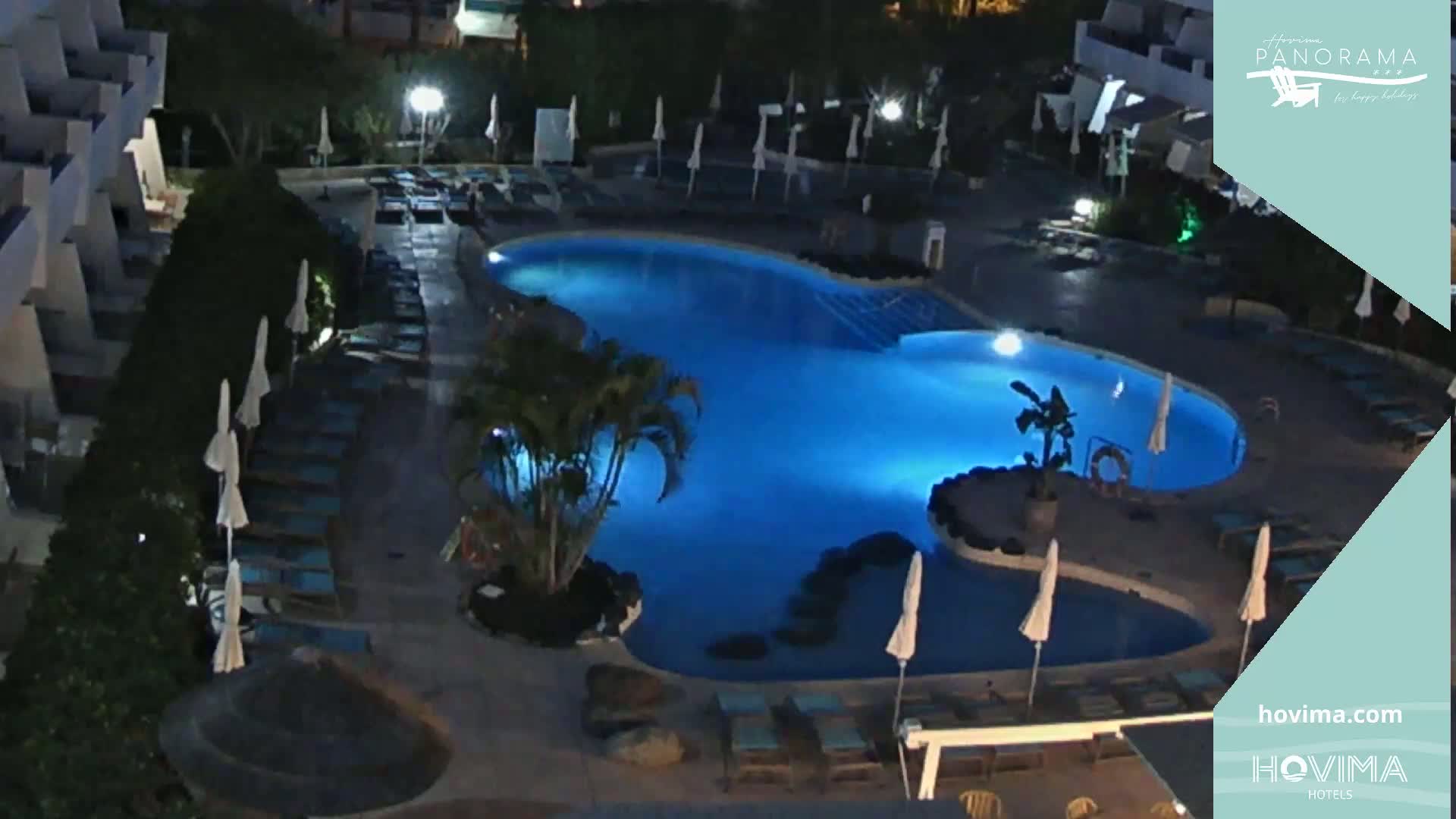 An illuminated, kidney-shaped swimming pool at night, surrounded by lounge chairs and umbrellas.