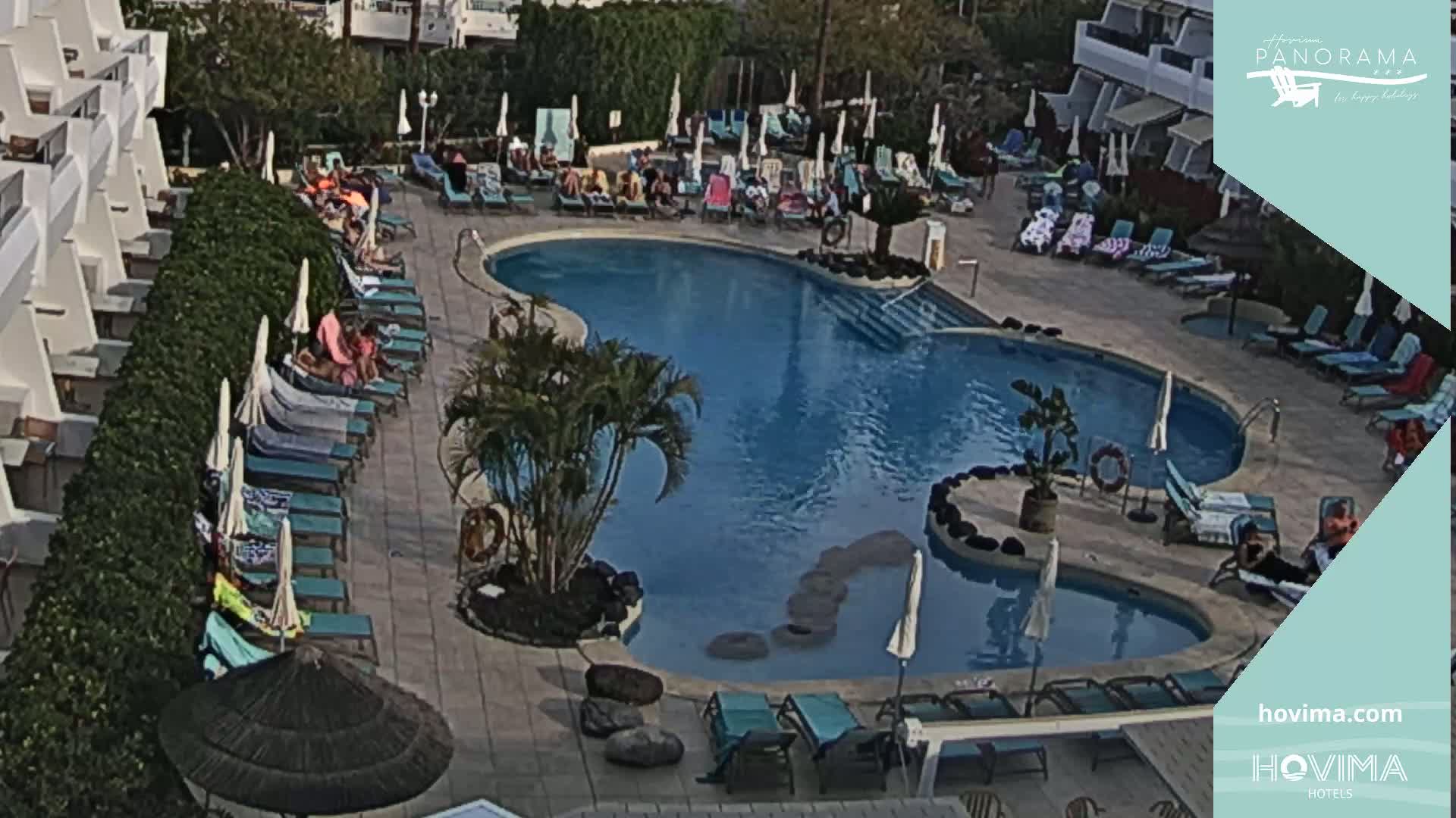 An aerial view shows a resort pool area with numerous lounge chairs surrounding a kidney-shaped pool under a mostly sunny sky.