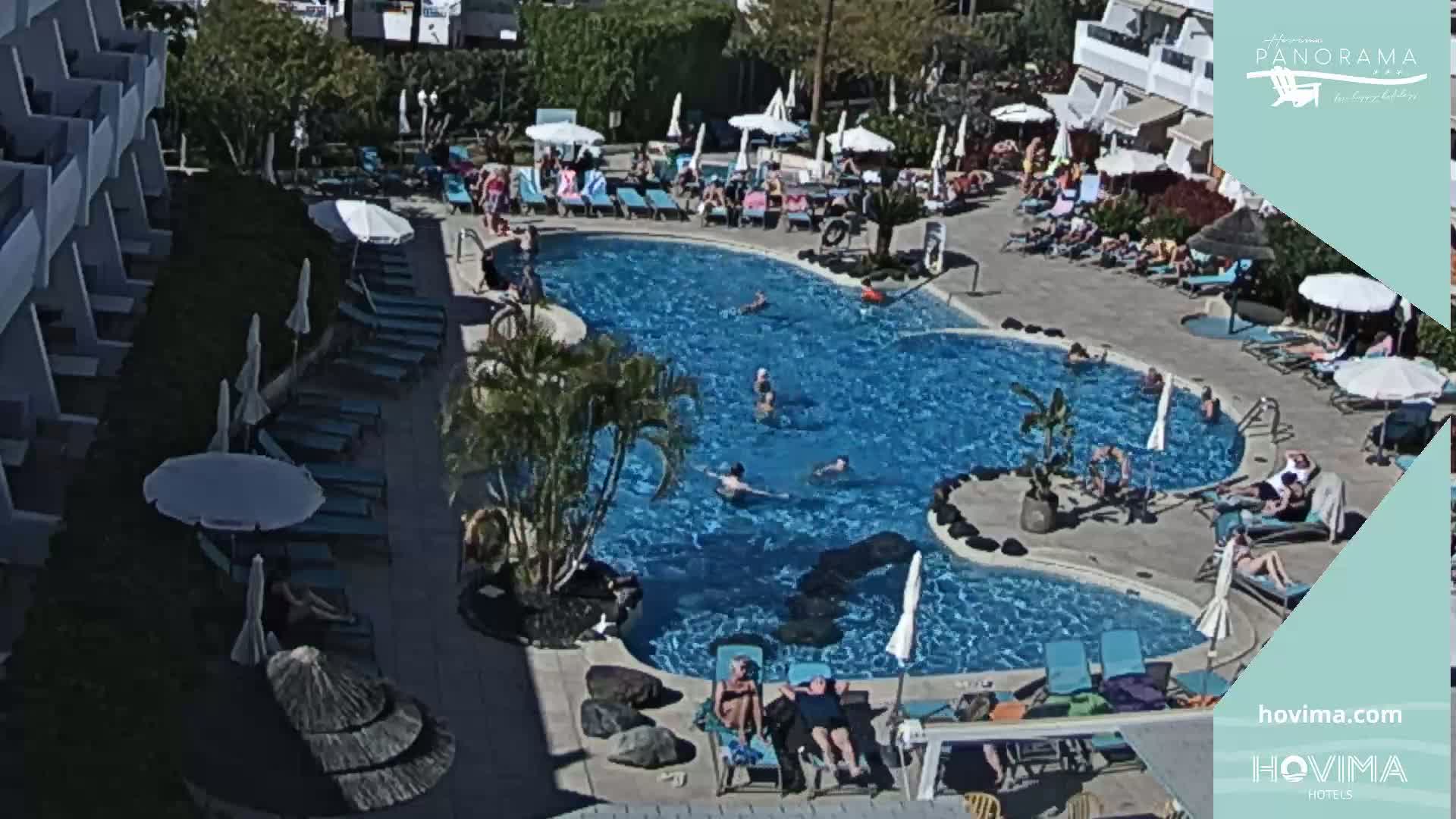 A sunny outdoor swimming pool surrounded by lounge chairs, umbrellas, and people swimming and sunbathing.