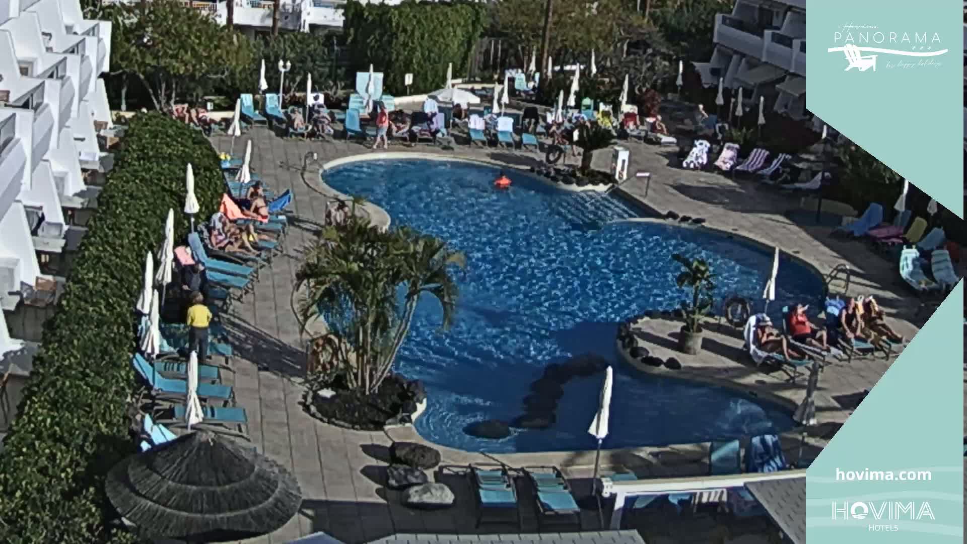 A sunny day at a resort pool area shows a kidney-shaped pool surrounded by lounge chairs and umbrellas, with many people relaxing around the pool.