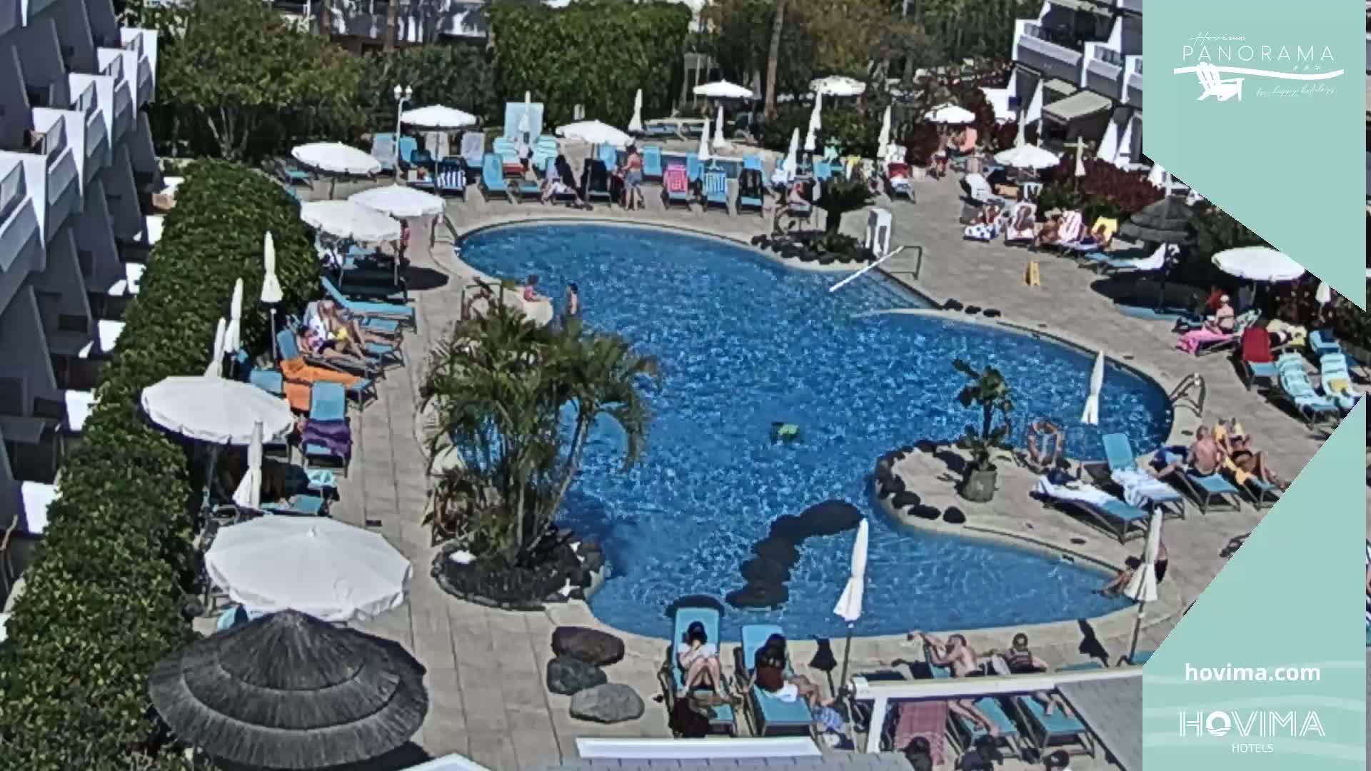 A resort pool area with many people lounging on chairs and under umbrellas on a sunny day.