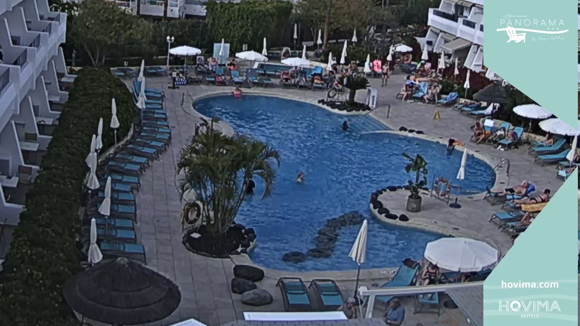 An outdoor pool area with numerous lounge chairs and umbrellas surrounding a kidney-shaped pool, where several people are swimming and sunbathing.