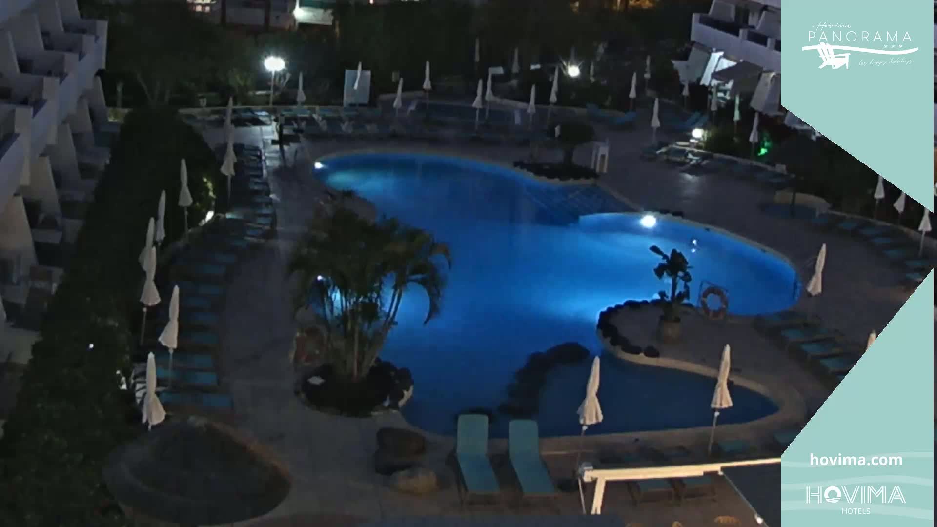 An illuminated, kidney-shaped swimming pool at night, surrounded by closed umbrellas and lounge chairs, next to a building.