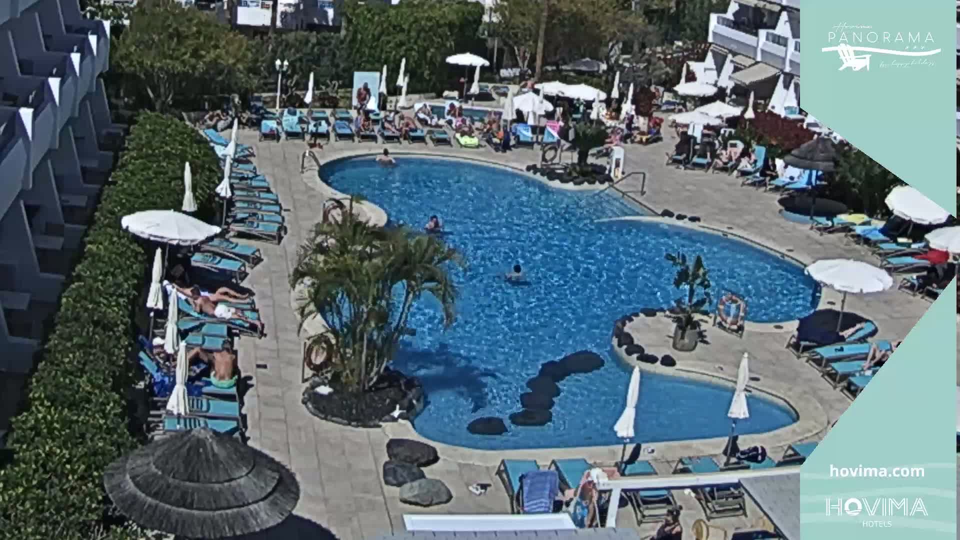 A sunny day at a resort pool area shows many people lounging on chairs and swimming in a kidney-shaped pool surrounded by palm trees and buildings.