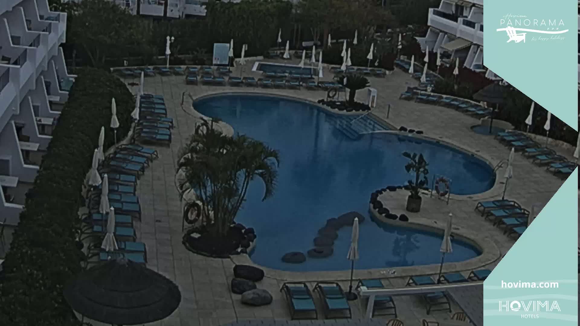 An overhead view of a resort pool area with many lounge chairs and umbrellas surrounding a kidney-shaped swimming pool on an overcast day.