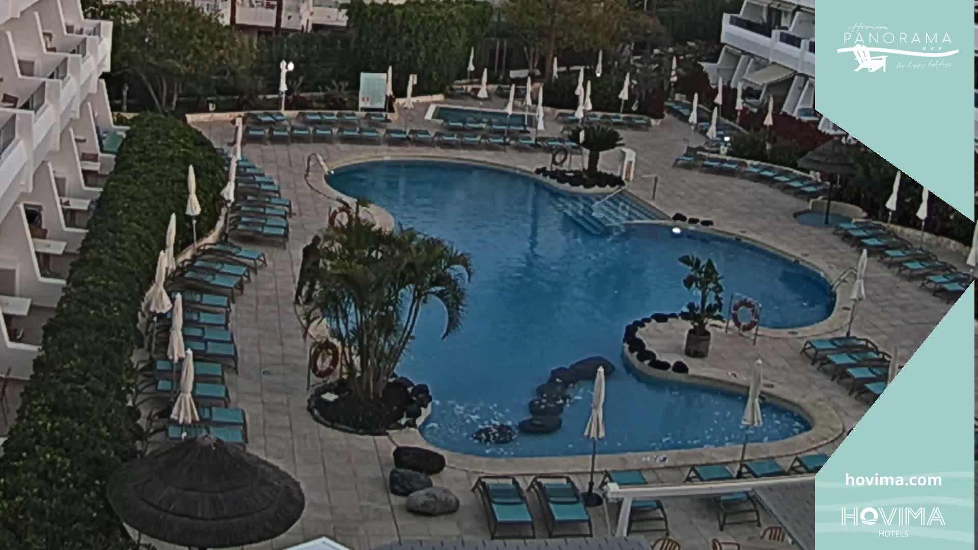 An aerial view shows a resort pool with surrounding lounge chairs and umbrellas under a partly cloudy sky.