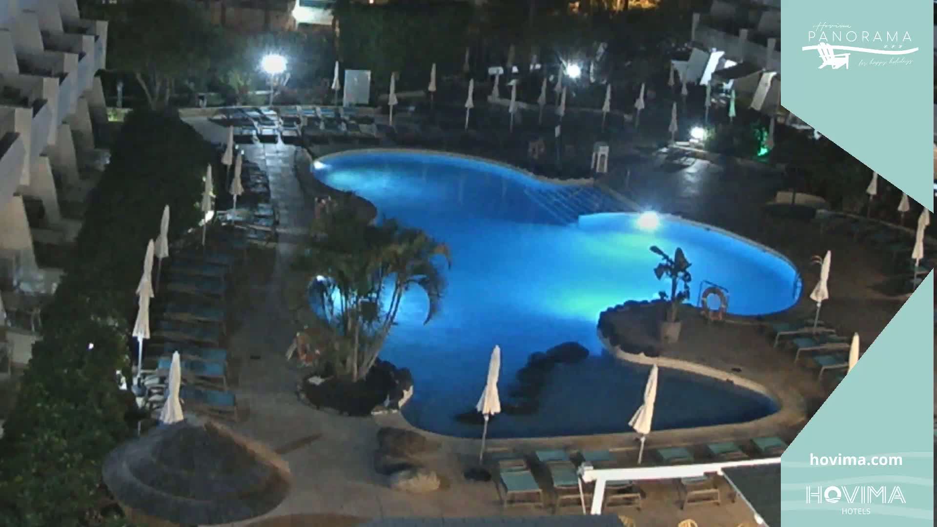 An illuminated swimming pool at night, surrounded by lounge chairs and umbrellas, next to a building.