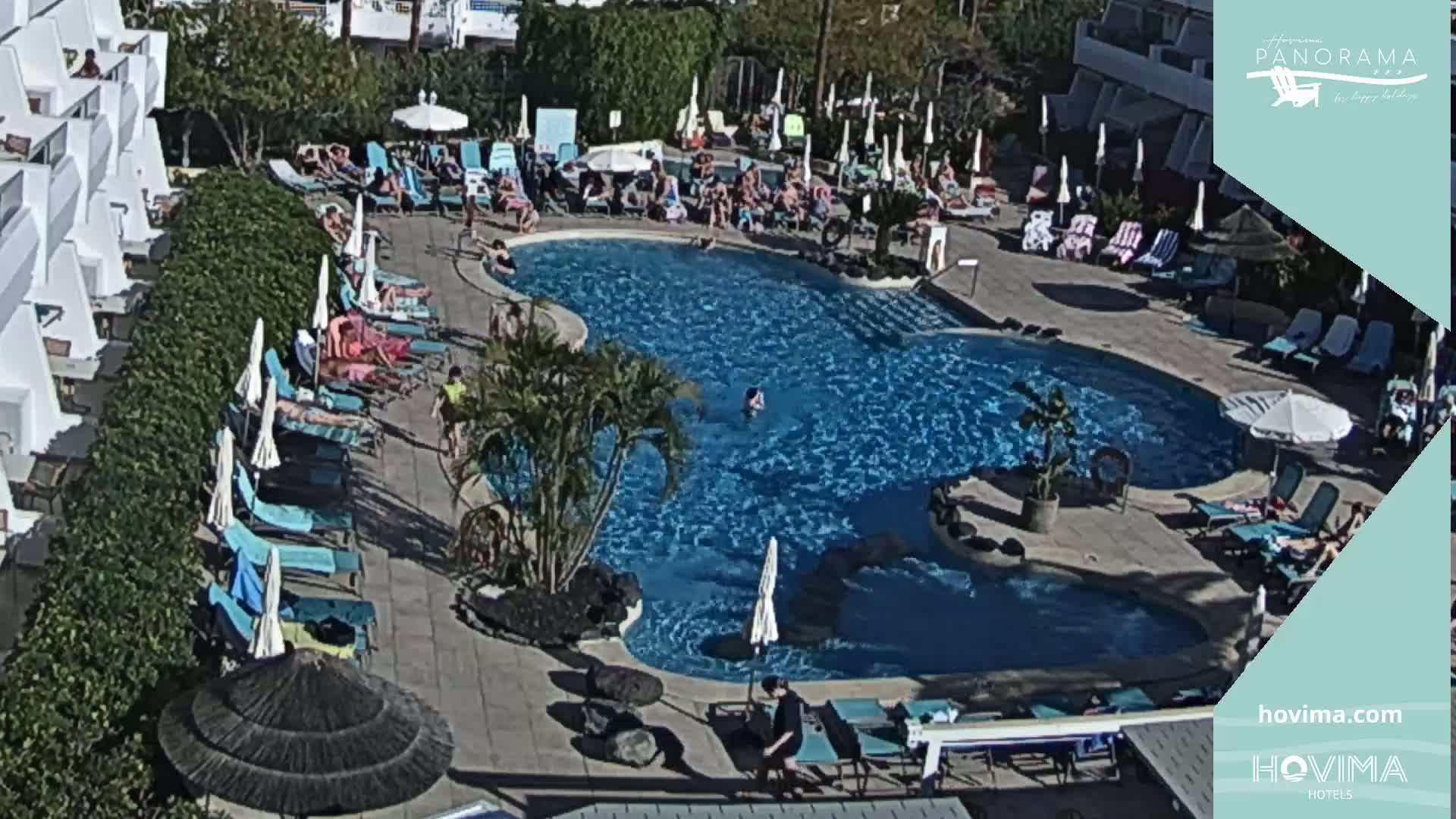 An outdoor pool with many people swimming and sunbathing on lounge chairs under umbrellas on a sunny day.