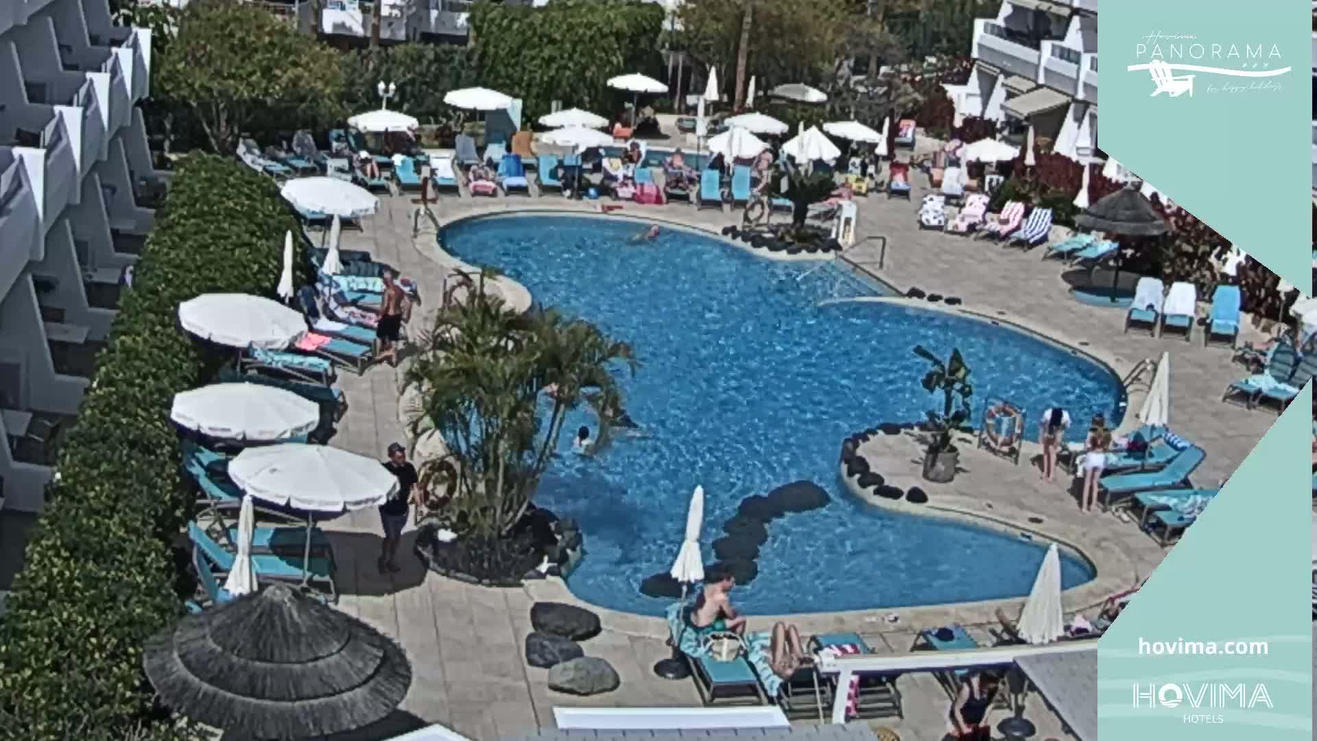 A sunny outdoor scene shows a kidney-shaped swimming pool surrounded by lounge chairs, umbrellas, and people relaxing, next to a multi-story building.