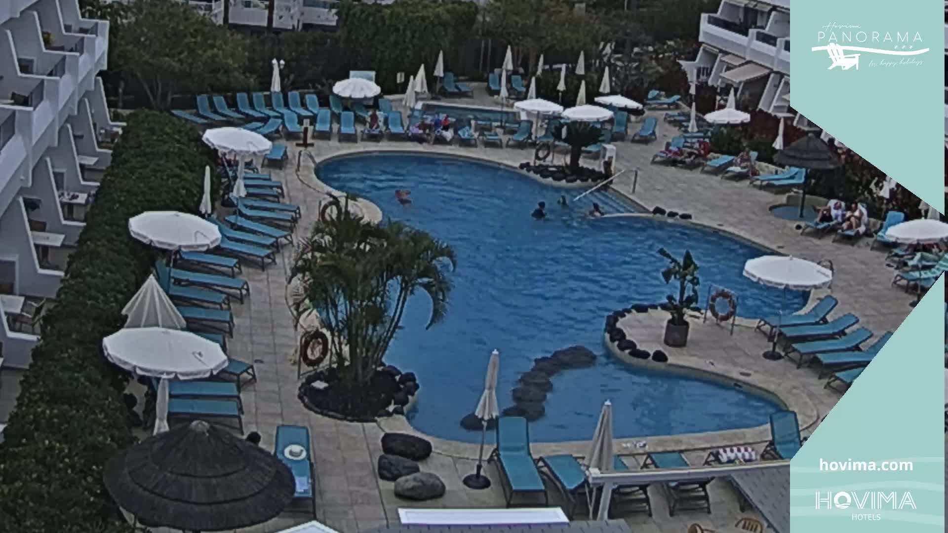 An outdoor swimming pool surrounded by lounge chairs and umbrellas, adjacent to a building, with several people in and around the pool.