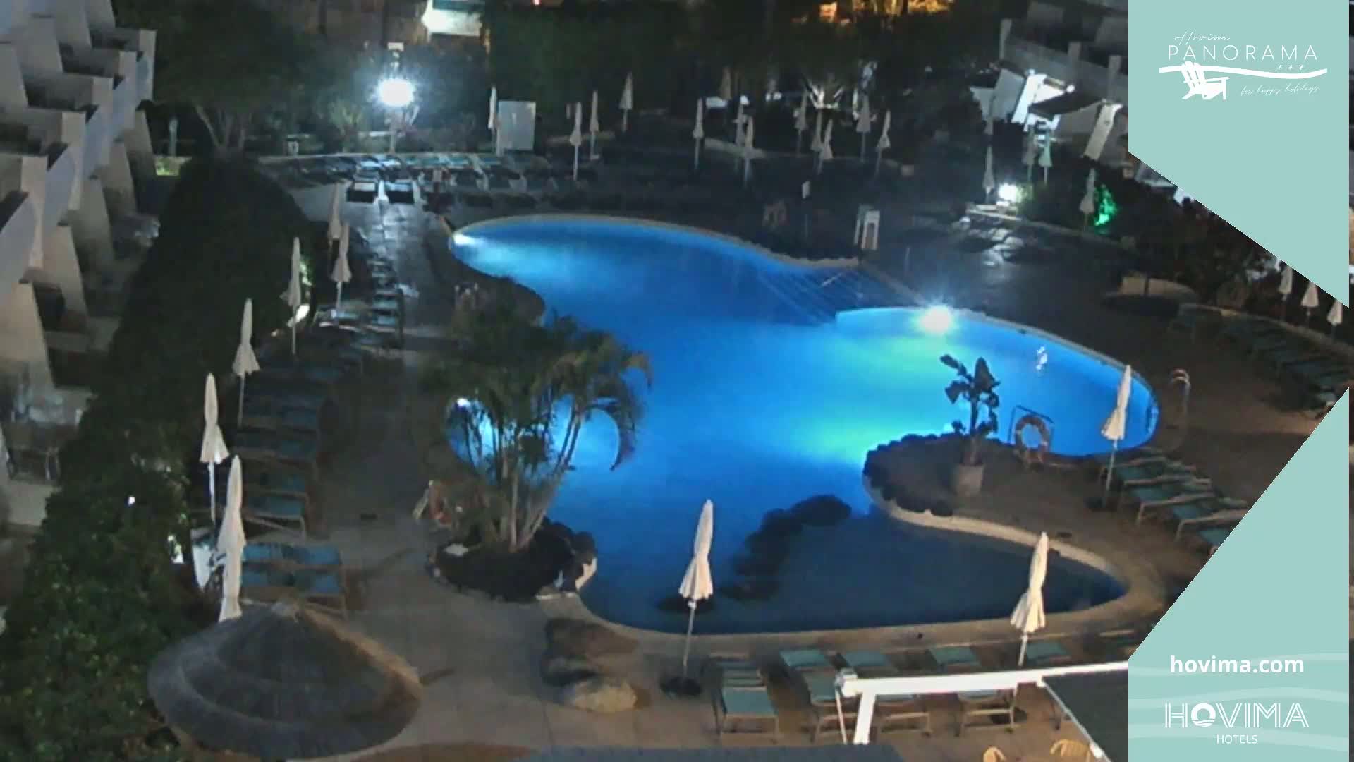 An illuminated, irregularly shaped swimming pool at night is surrounded by lounge chairs and umbrellas.