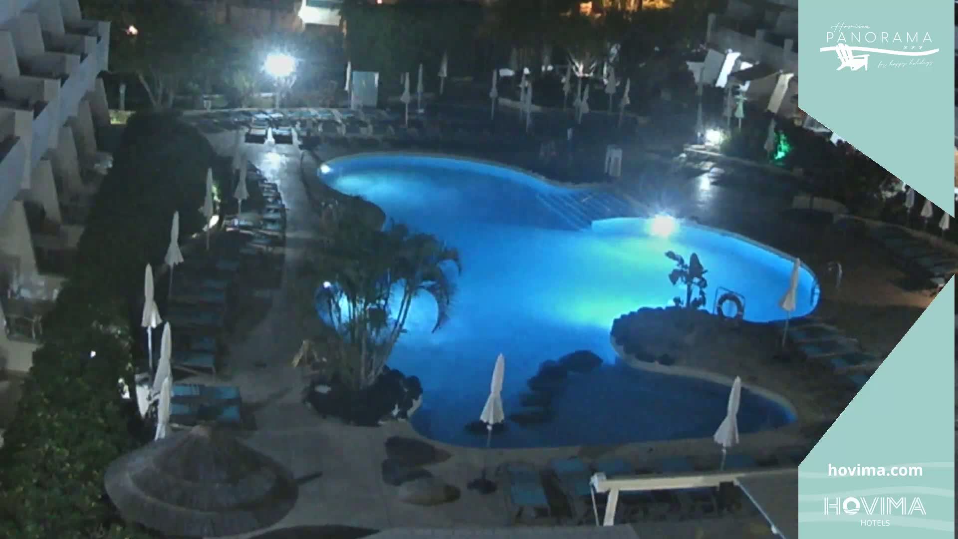 An illuminated, kidney-shaped swimming pool at night, surrounded by closed lounge chairs and umbrellas.