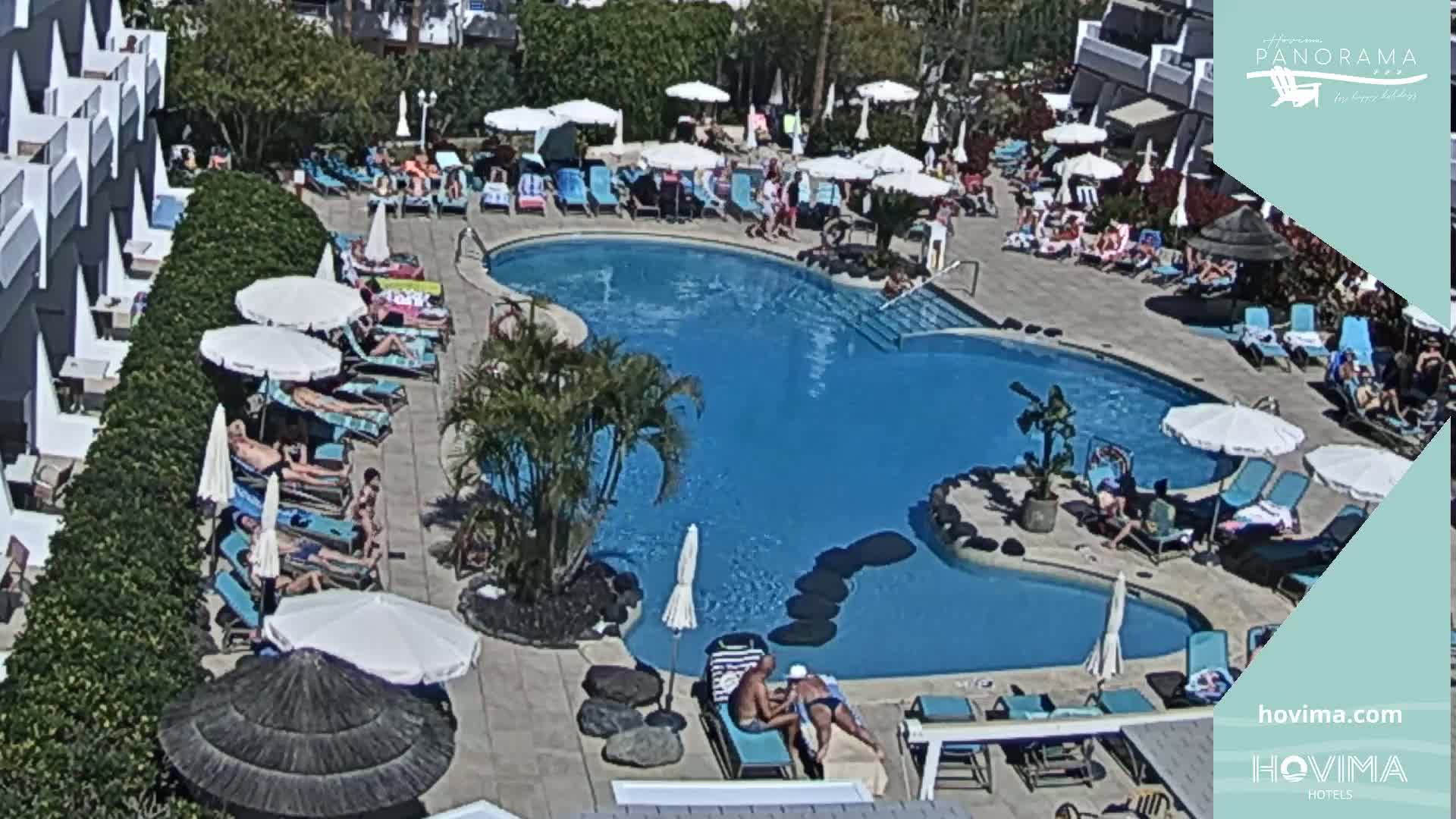 A resort pool area with many people relaxing on lounge chairs under umbrellas on a sunny day.