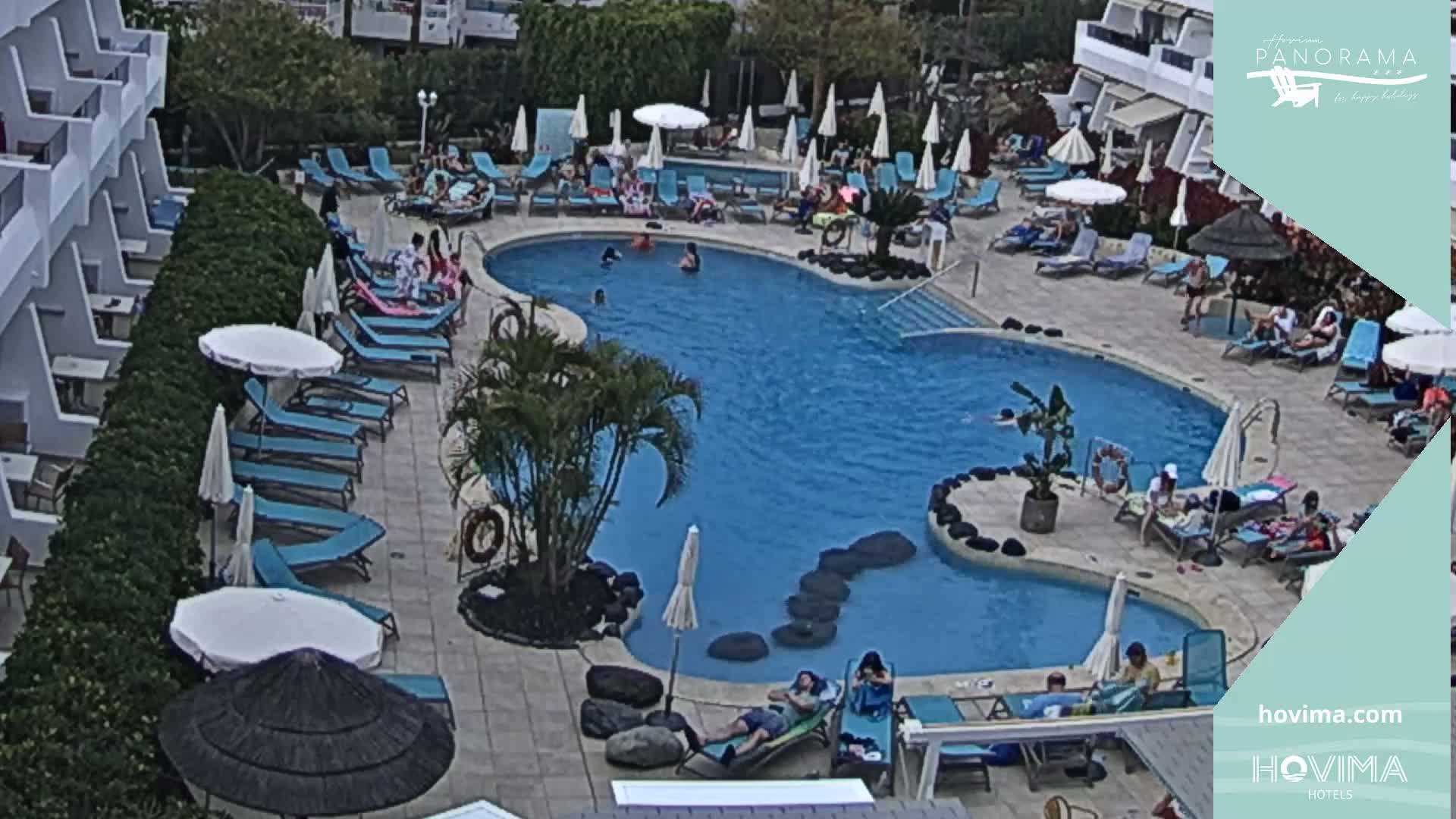 An outdoor pool surrounded by lounge chairs and umbrellas, with people swimming and relaxing in mostly sunny weather.