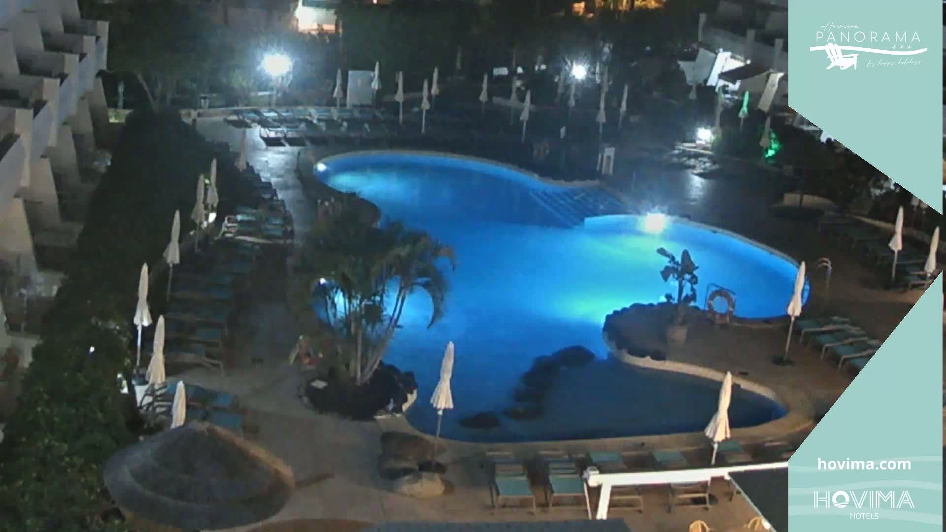 An illuminated swimming pool at night, surrounded by lounge chairs and umbrellas, is situated next to a building.