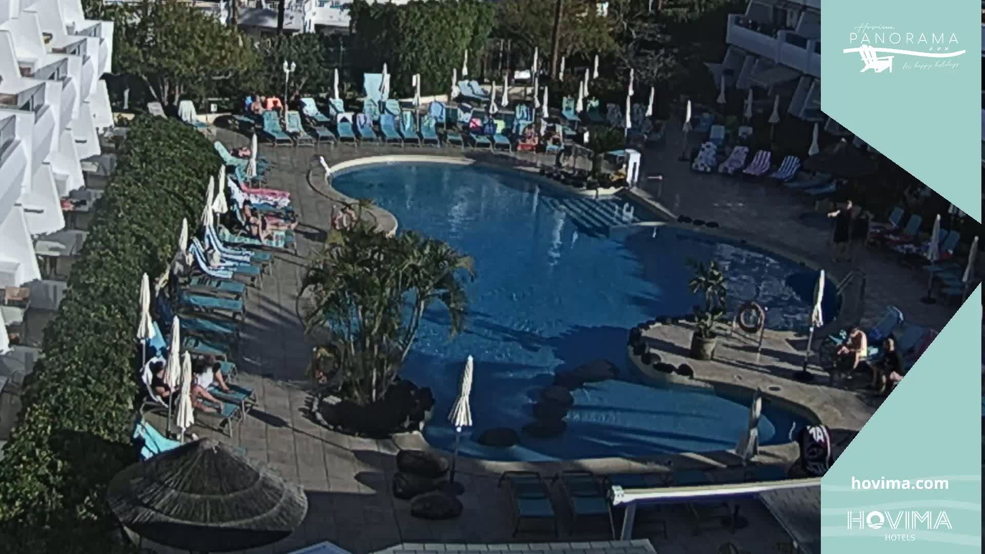 An outdoor pool area with numerous lounge chairs and umbrellas surrounding a kidney-shaped pool, populated by several people, under sunny conditions.