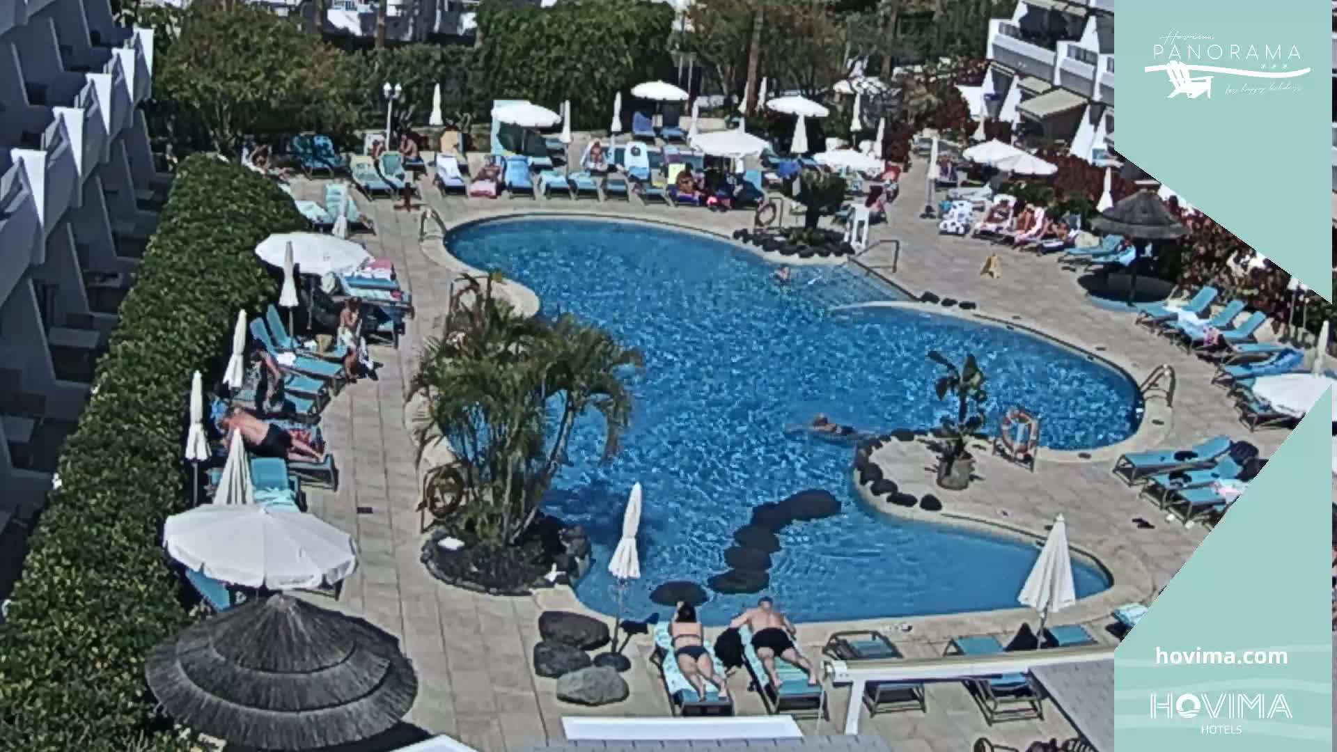 An outdoor swimming pool surrounded by lounge chairs and umbrellas, with people swimming and sunbathing on a sunny day.