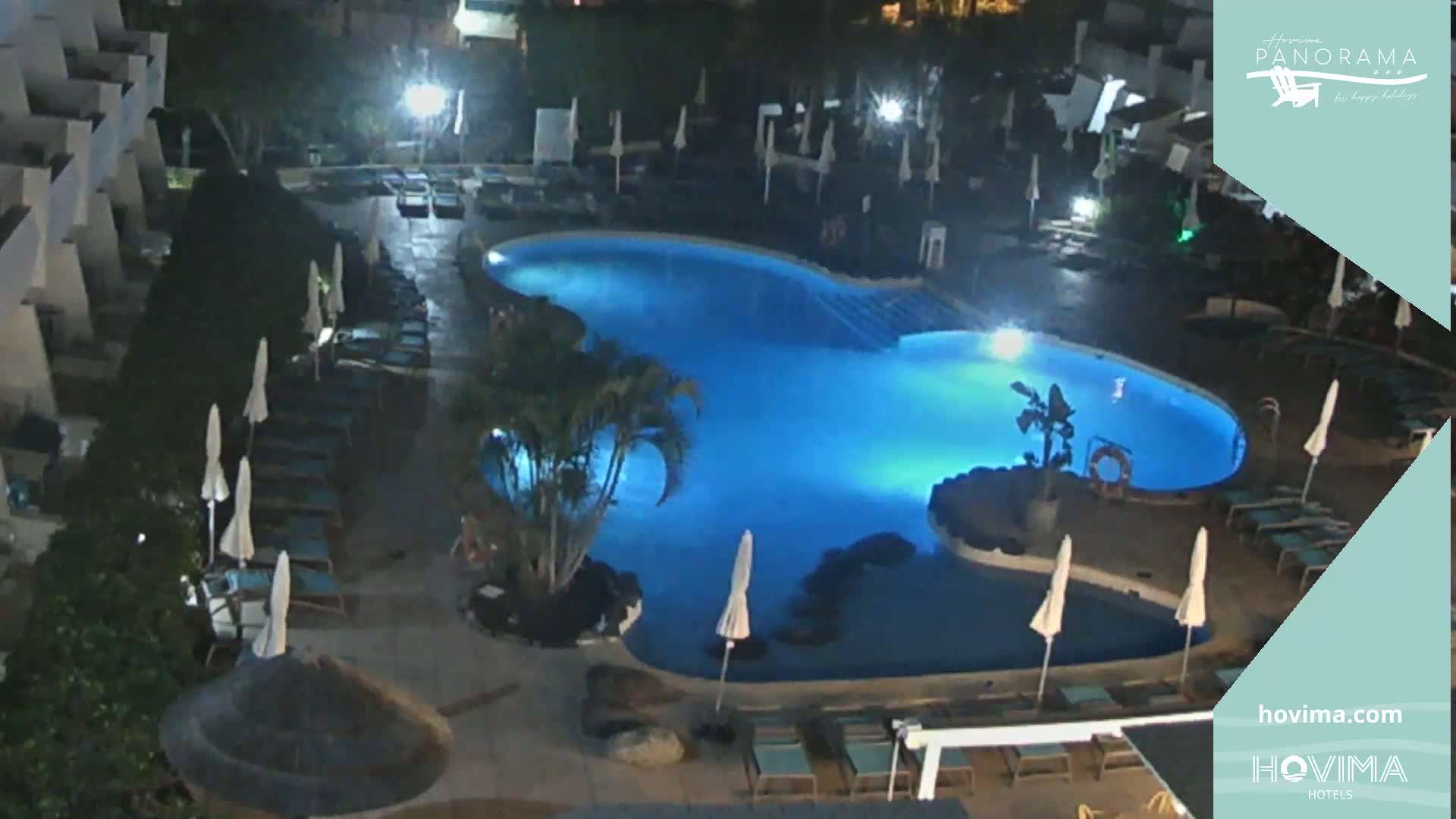 An illuminated, kidney-shaped swimming pool at night, surrounded by lounge chairs and umbrellas.