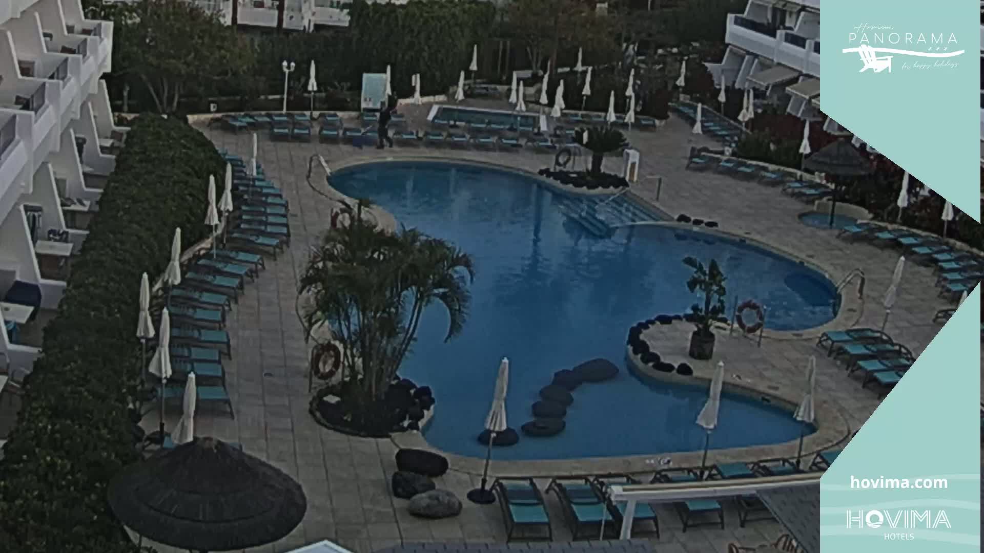 An aerial view shows a resort pool with surrounding lounge chairs and umbrellas under a partly cloudy sky.