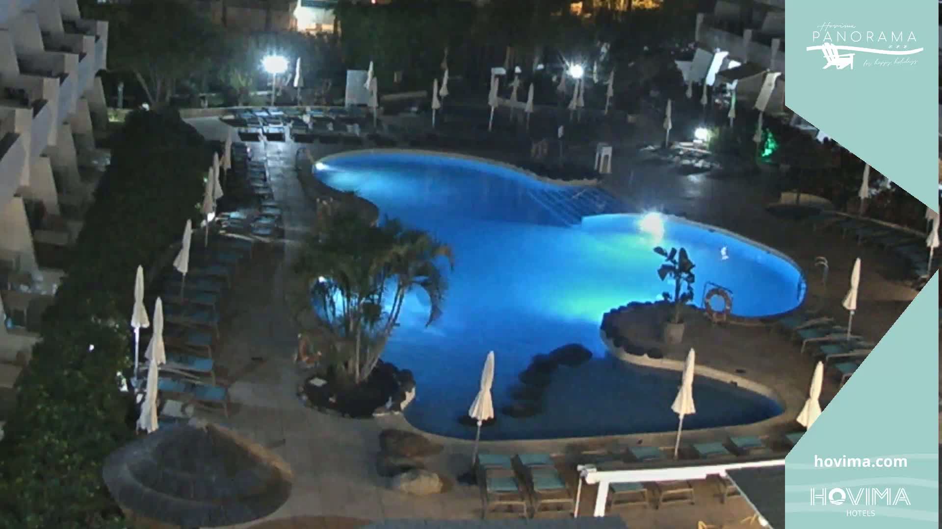 A nighttime, overhead view of a resort pool area with lounge chairs and umbrellas surrounding a brightly lit, kidney-shaped pool.