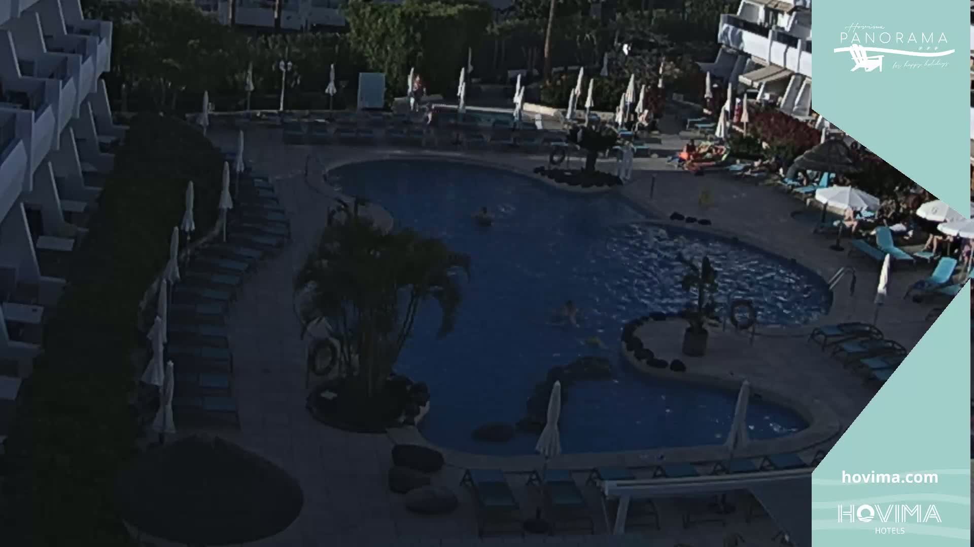 An outdoor swimming pool surrounded by lounge chairs and umbrellas, with a hotel building in the background under partly cloudy conditions.