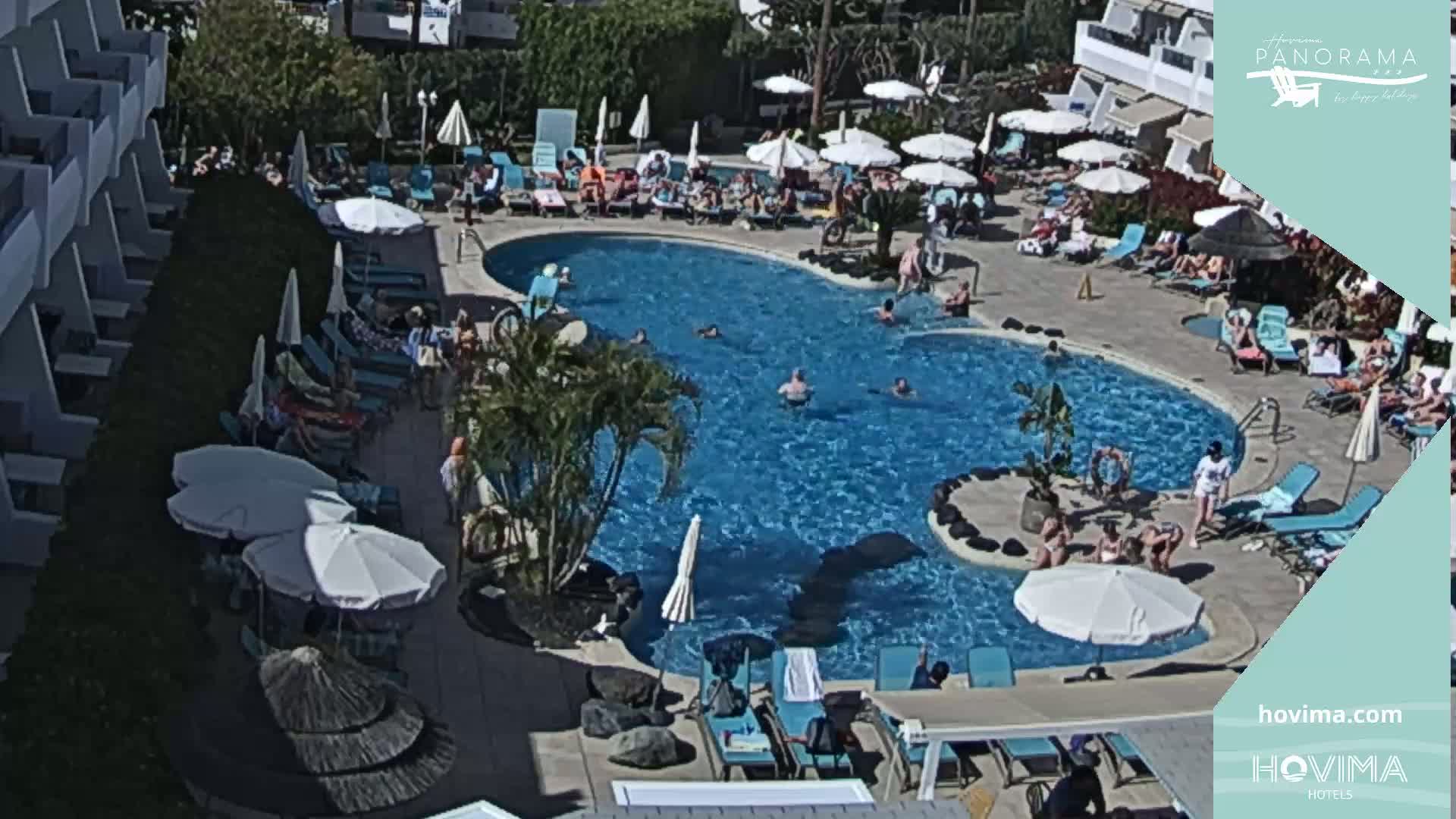 A sunny outdoor pool area with many people swimming and lounging on chairs under umbrellas.