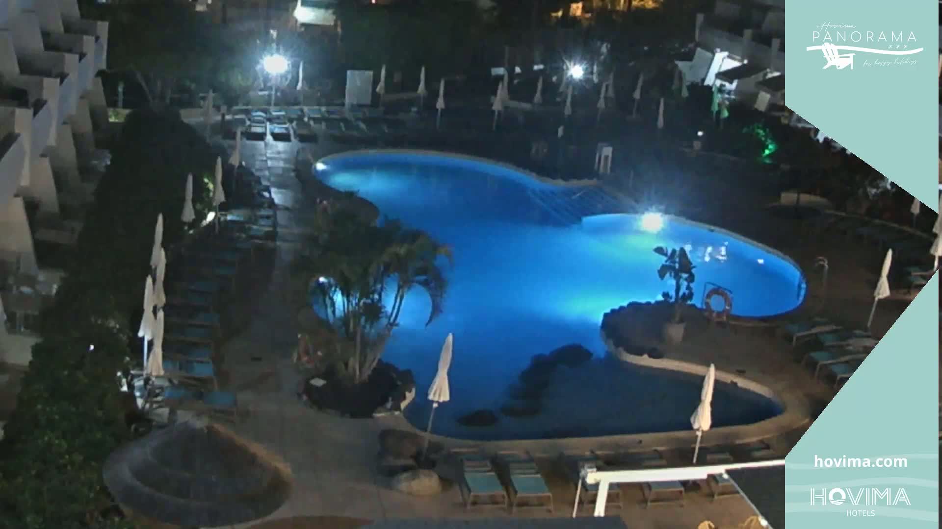 An illuminated, irregularly shaped swimming pool at night, surrounded by lounge chairs and umbrellas, is situated next to a multi-story building.