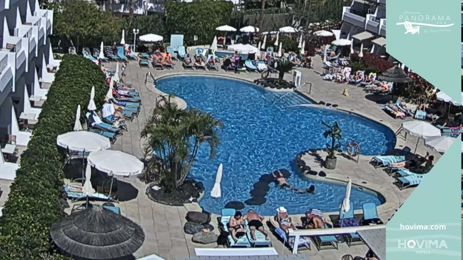 An outdoor pool area with many people lounging on chairs and in the water under a sunny sky.