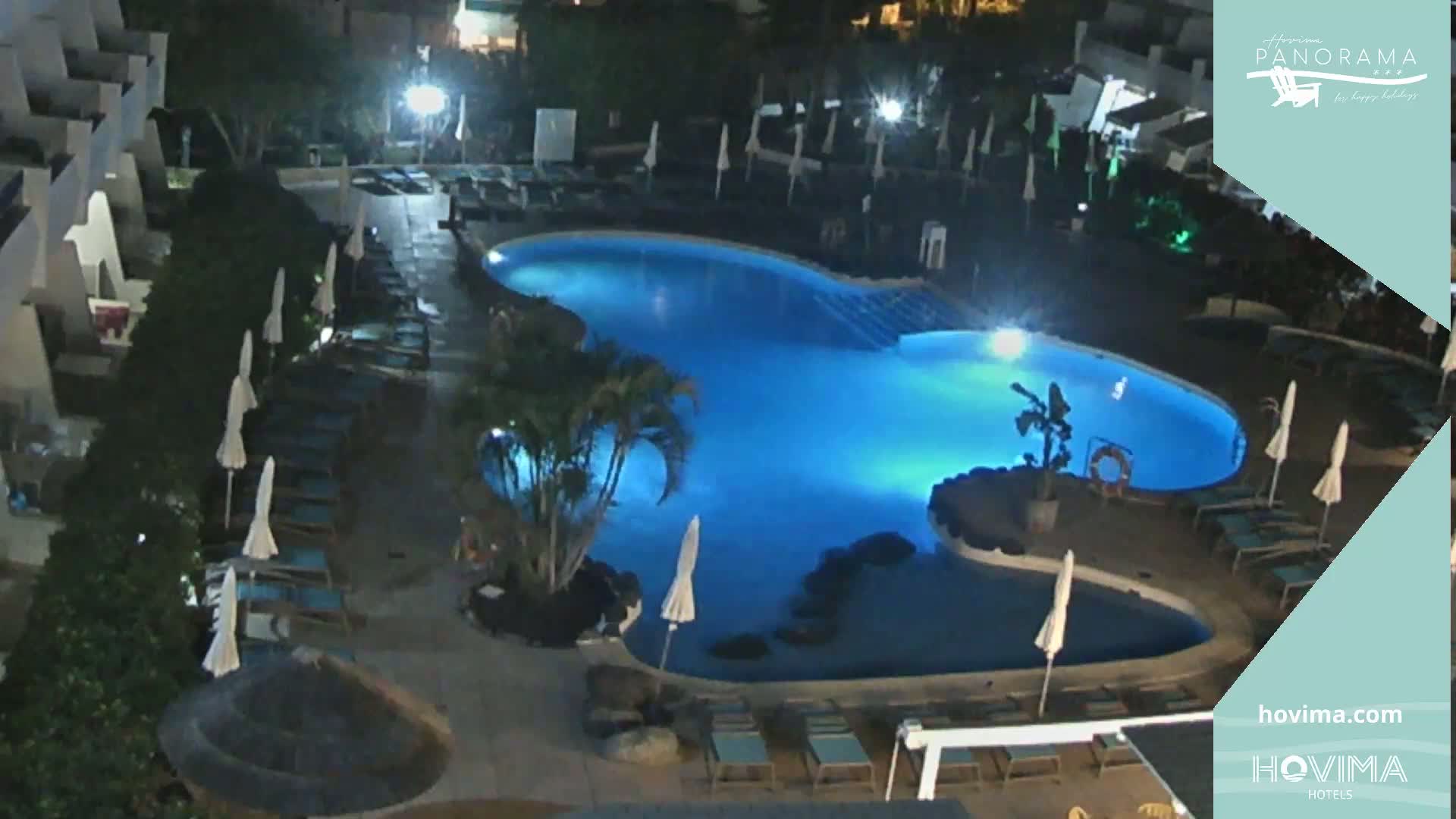 A nighttime aerial view shows a kidney-shaped swimming pool illuminated in blue, surrounded by lounge chairs and palm trees.