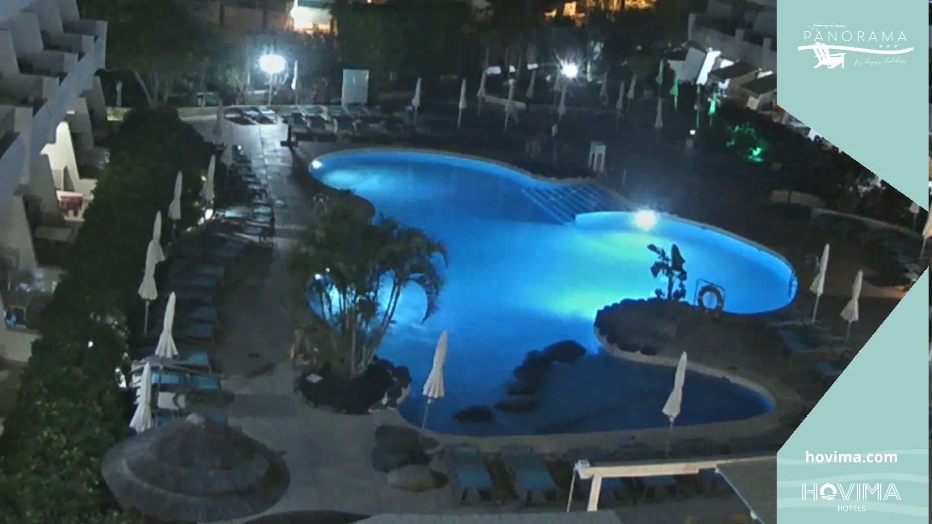 A brightly lit, kidney-shaped swimming pool at night, surrounded by lounge chairs and palm trees.