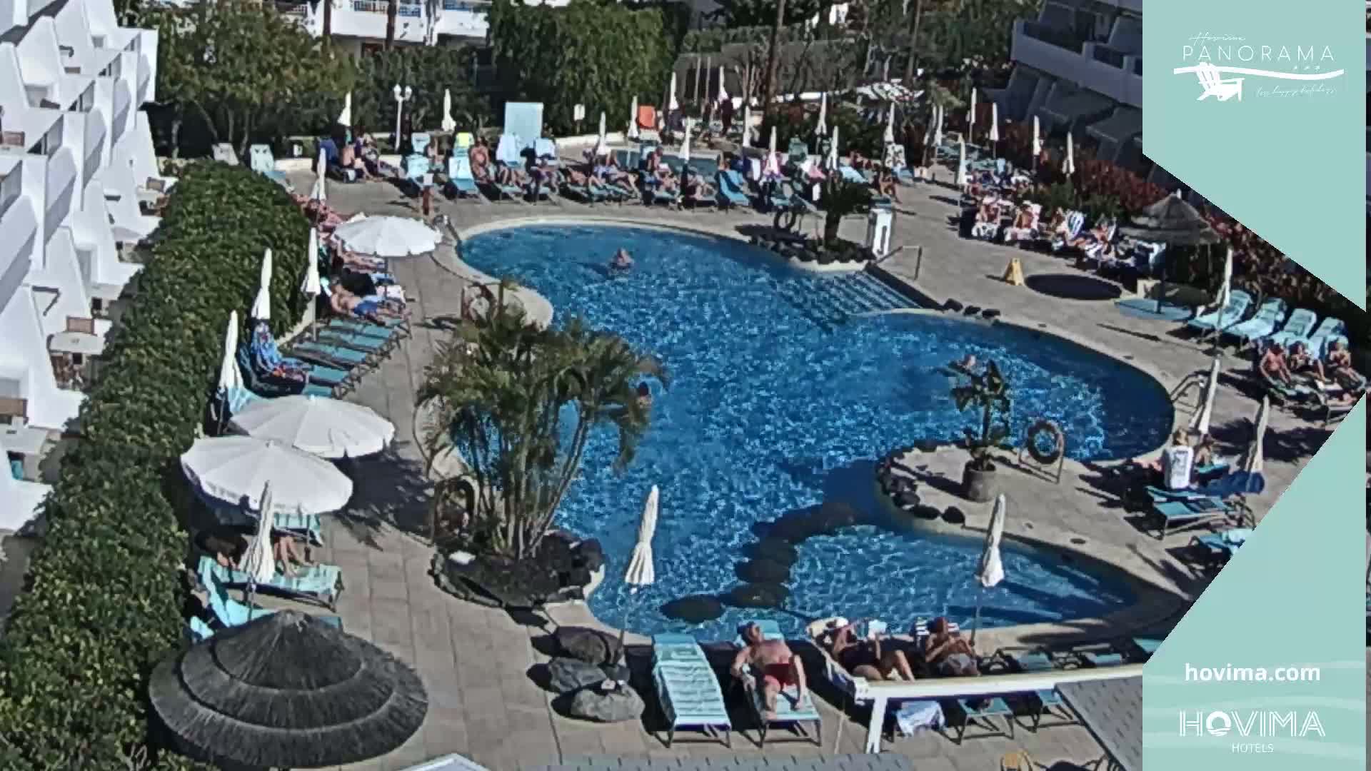 A resort pool area with many people sunbathing and swimming under a sunny sky.