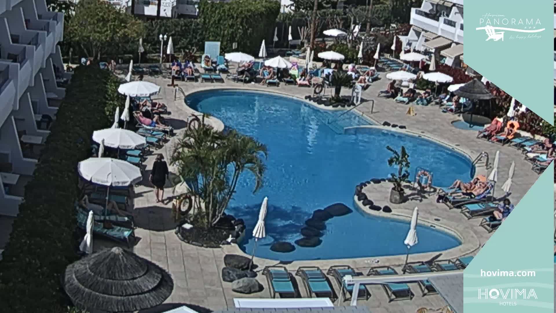 A resort pool area with numerous people sunbathing and swimming under a sunny sky.