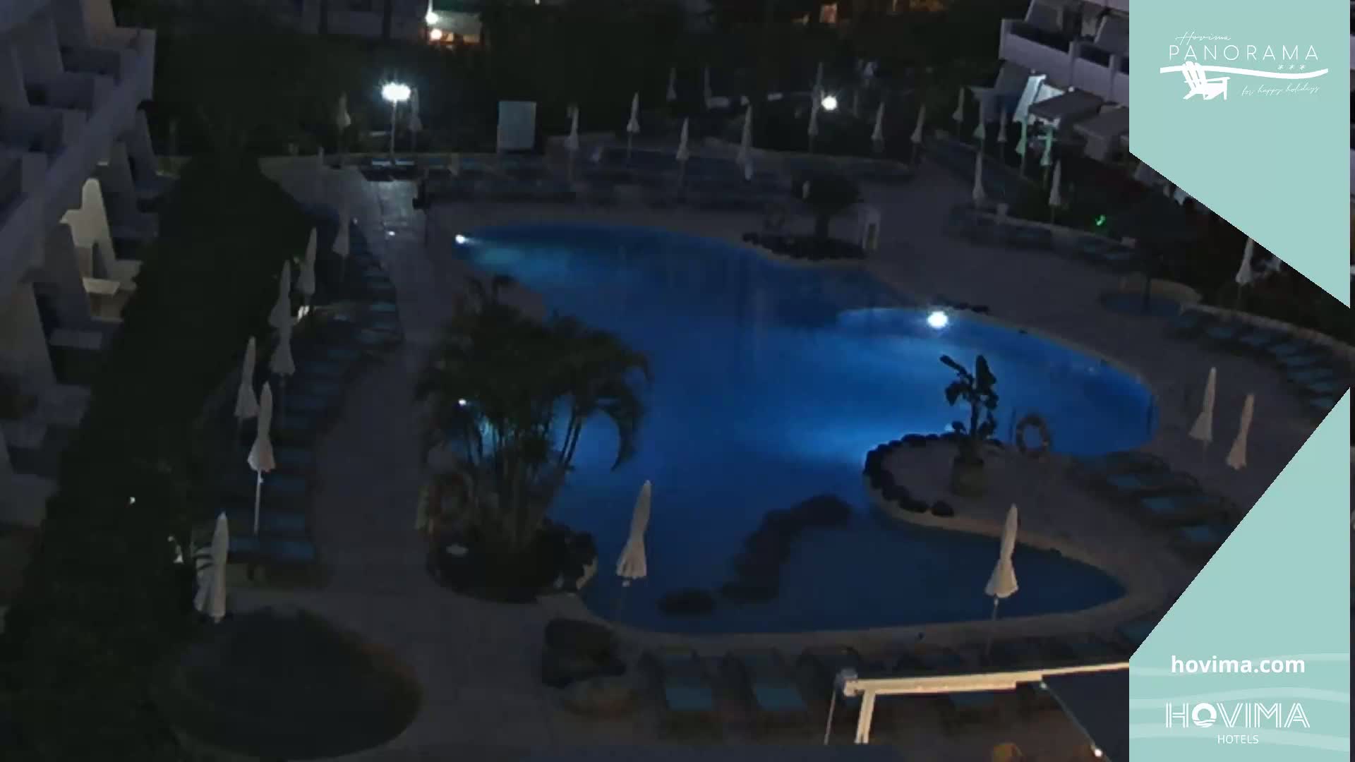 An illuminated, irregularly shaped swimming pool at night is surrounded by lounge chairs and umbrellas.