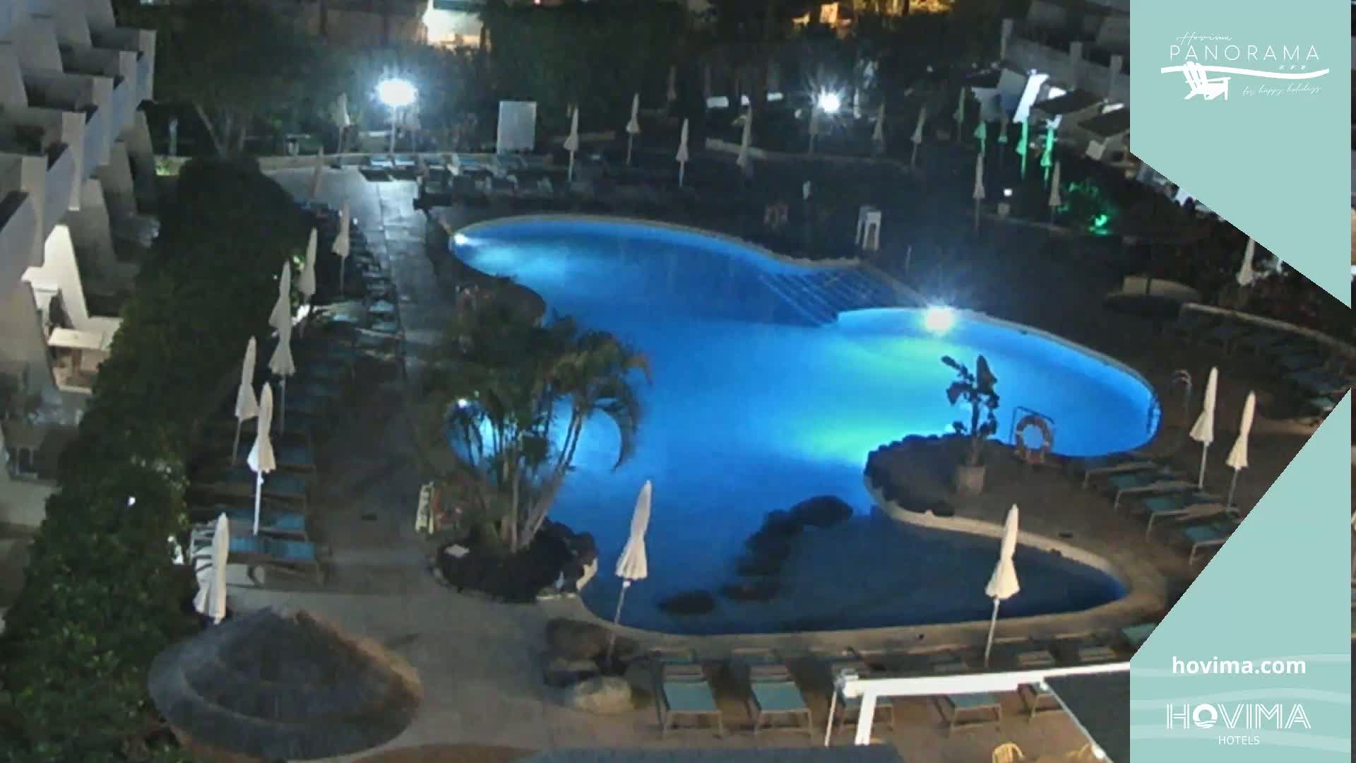 An illuminated, kidney-shaped swimming pool is surrounded by lounge chairs and umbrellas at night.