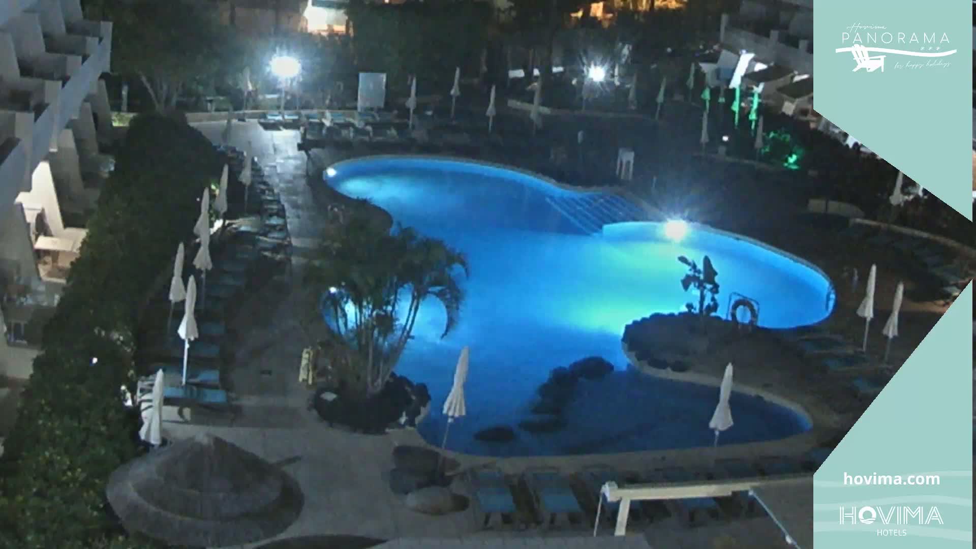 A brightly lit, kidney-shaped swimming pool at night, surrounded by closed umbrellas and lounge chairs, is nestled within a resort complex.
