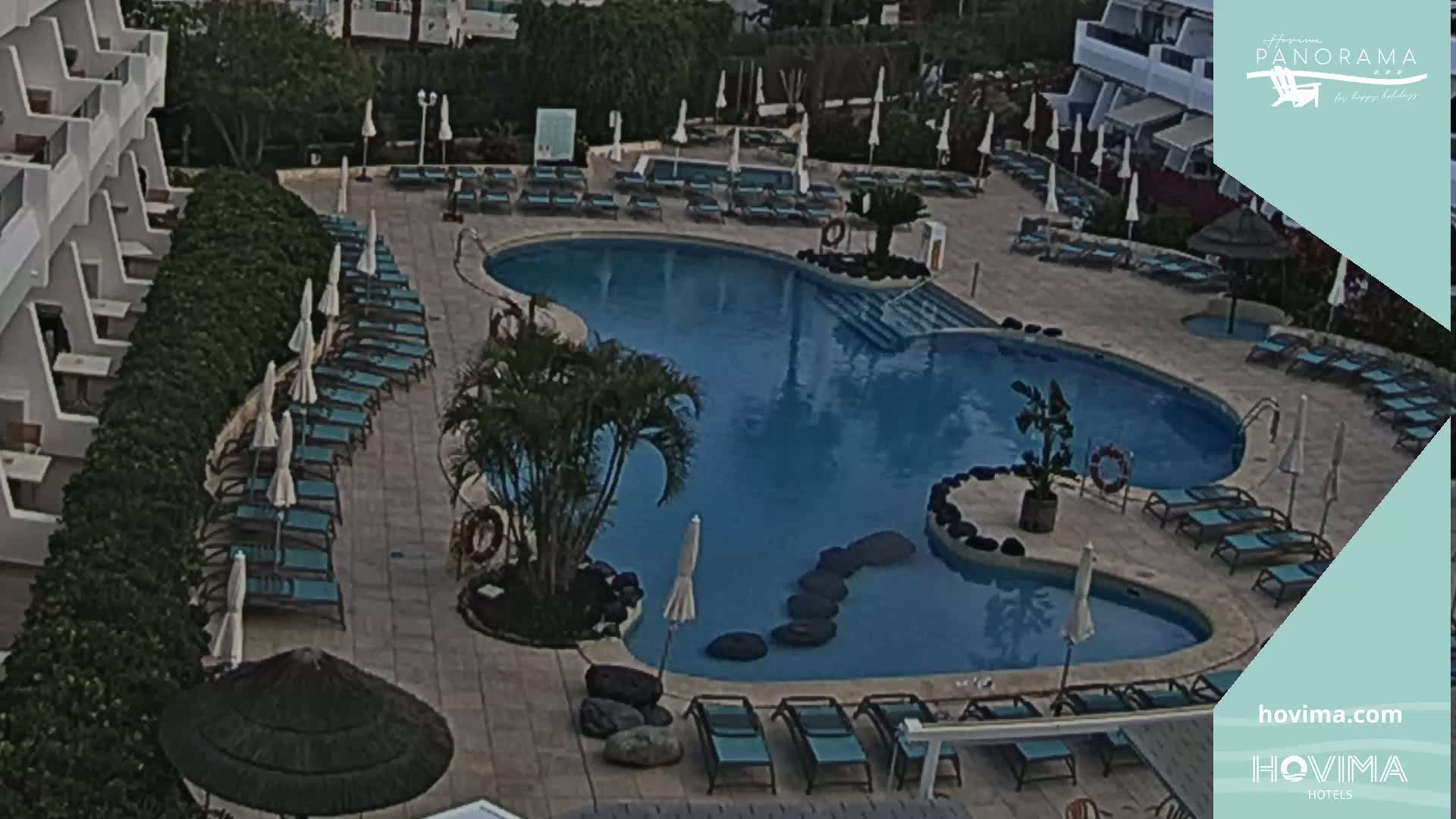 An outdoor swimming pool with surrounding lounge chairs and umbrellas sits next to a building under a mostly clear sky.