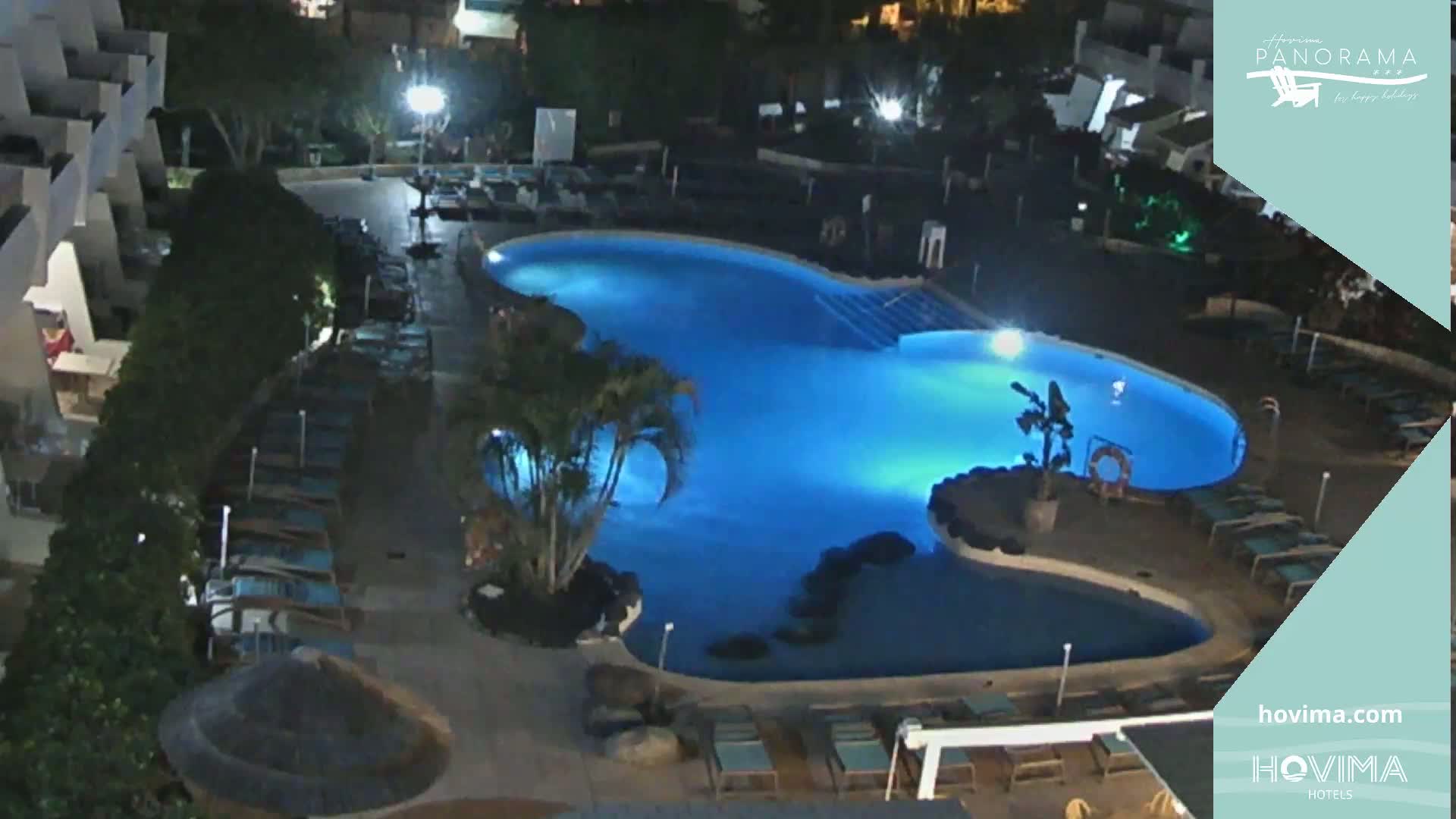 A nighttime aerial view of a resort pool area, illuminated by artificial lights, with surrounding buildings and lounge chairs.