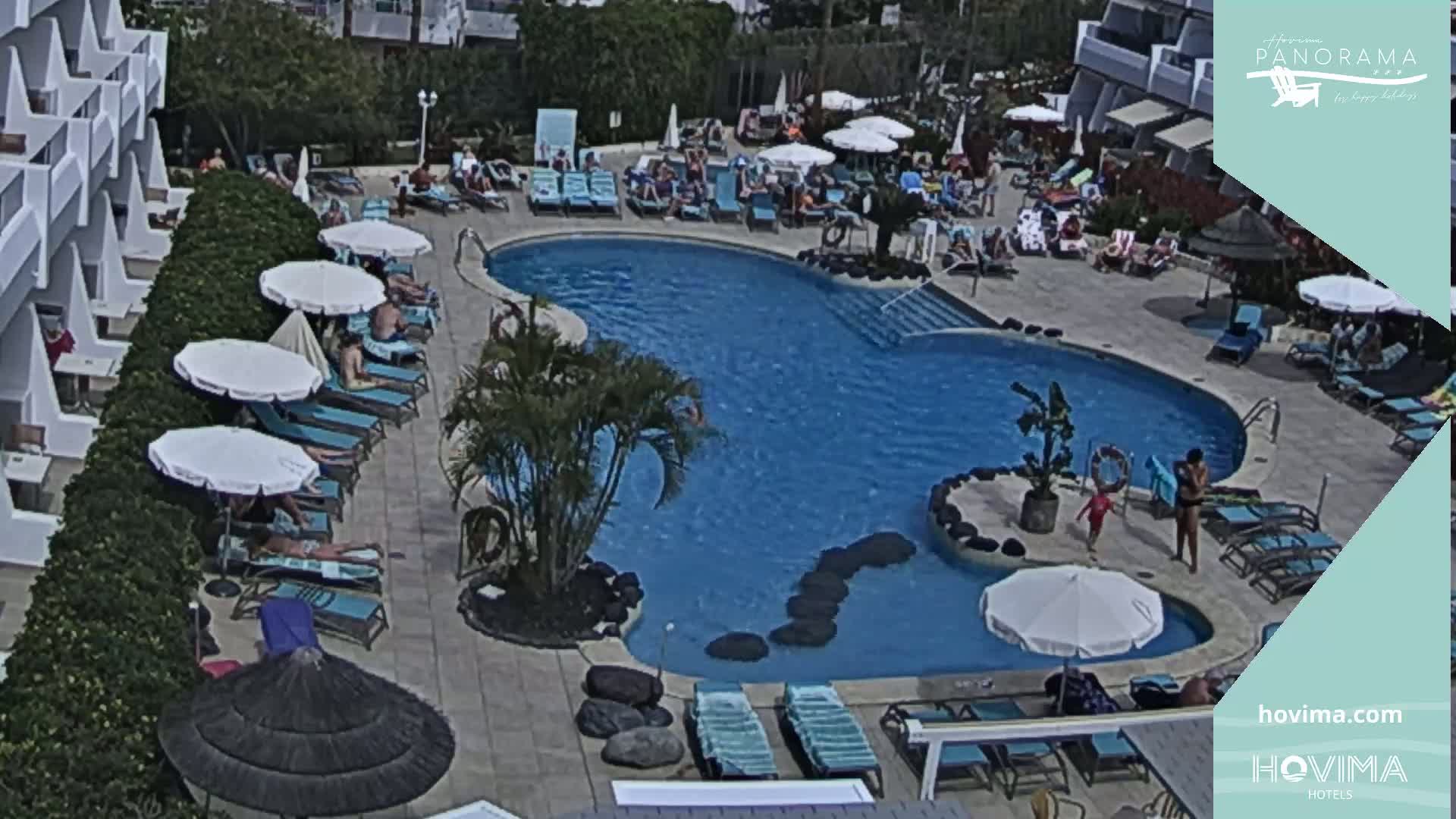 An outdoor swimming pool surrounded by lounge chairs and umbrellas, with people relaxing in the sun on a sunny day.