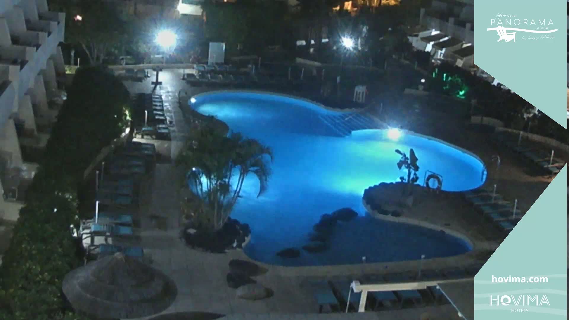 An illuminated swimming pool at night, surrounded by lounge chairs and vegetation, is seen from an elevated vantage point.