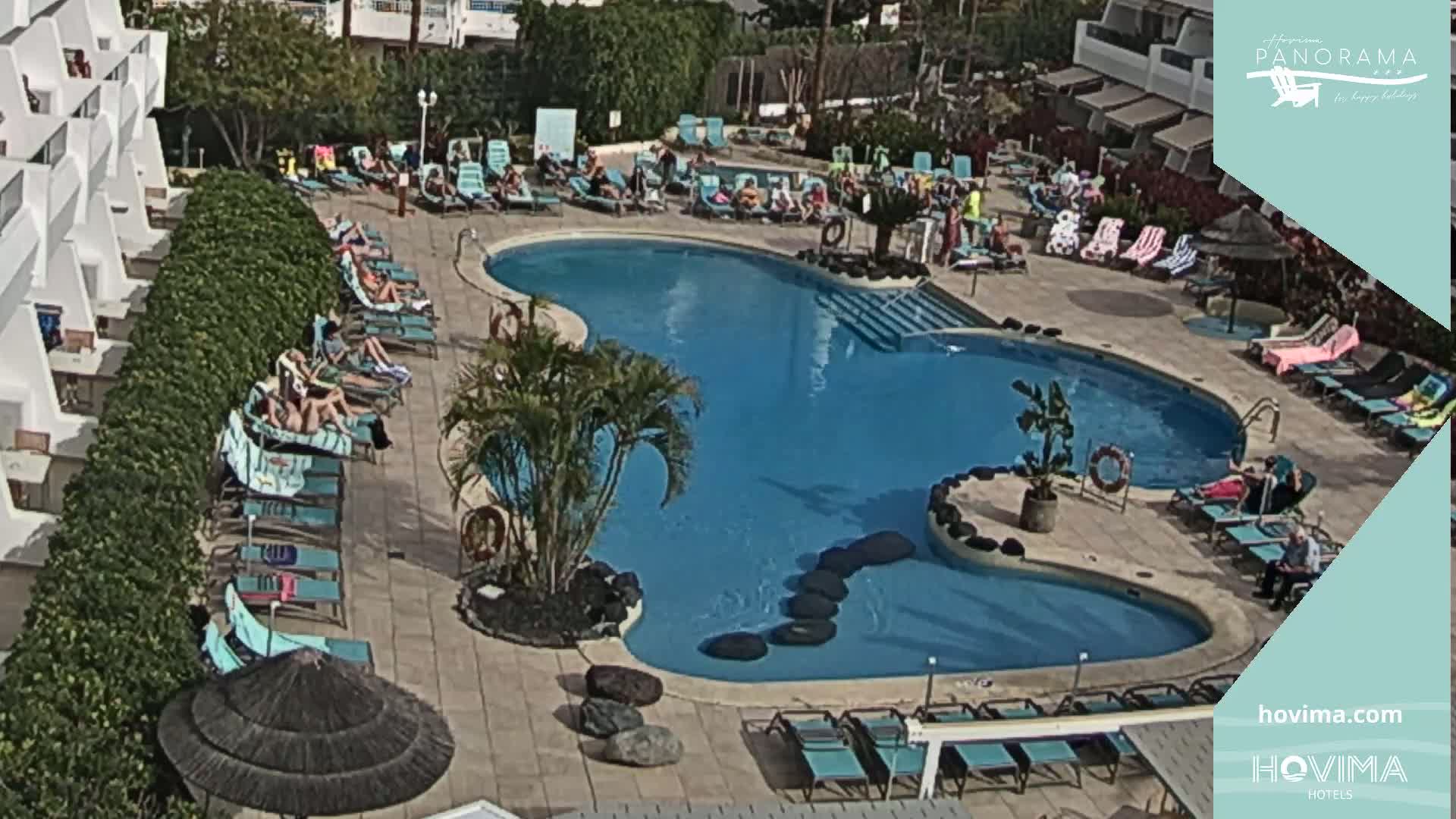 A sunny outdoor scene shows a kidney-shaped swimming pool surrounded by lounge chairs and people relaxing, with a row of buildings visible in the background.