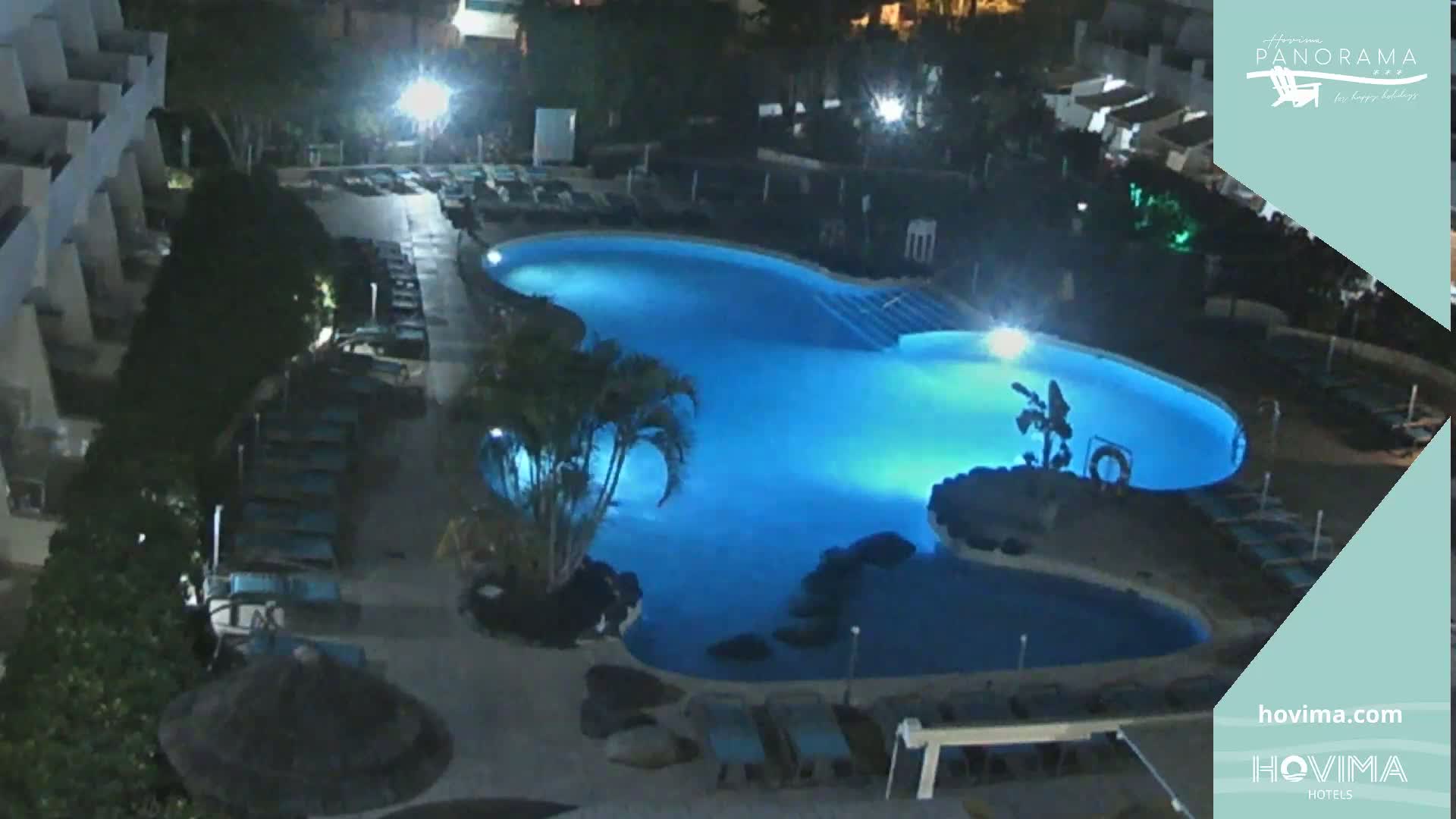 A brightly lit, kidney-shaped swimming pool at night, surrounded by lounge chairs and palm trees.