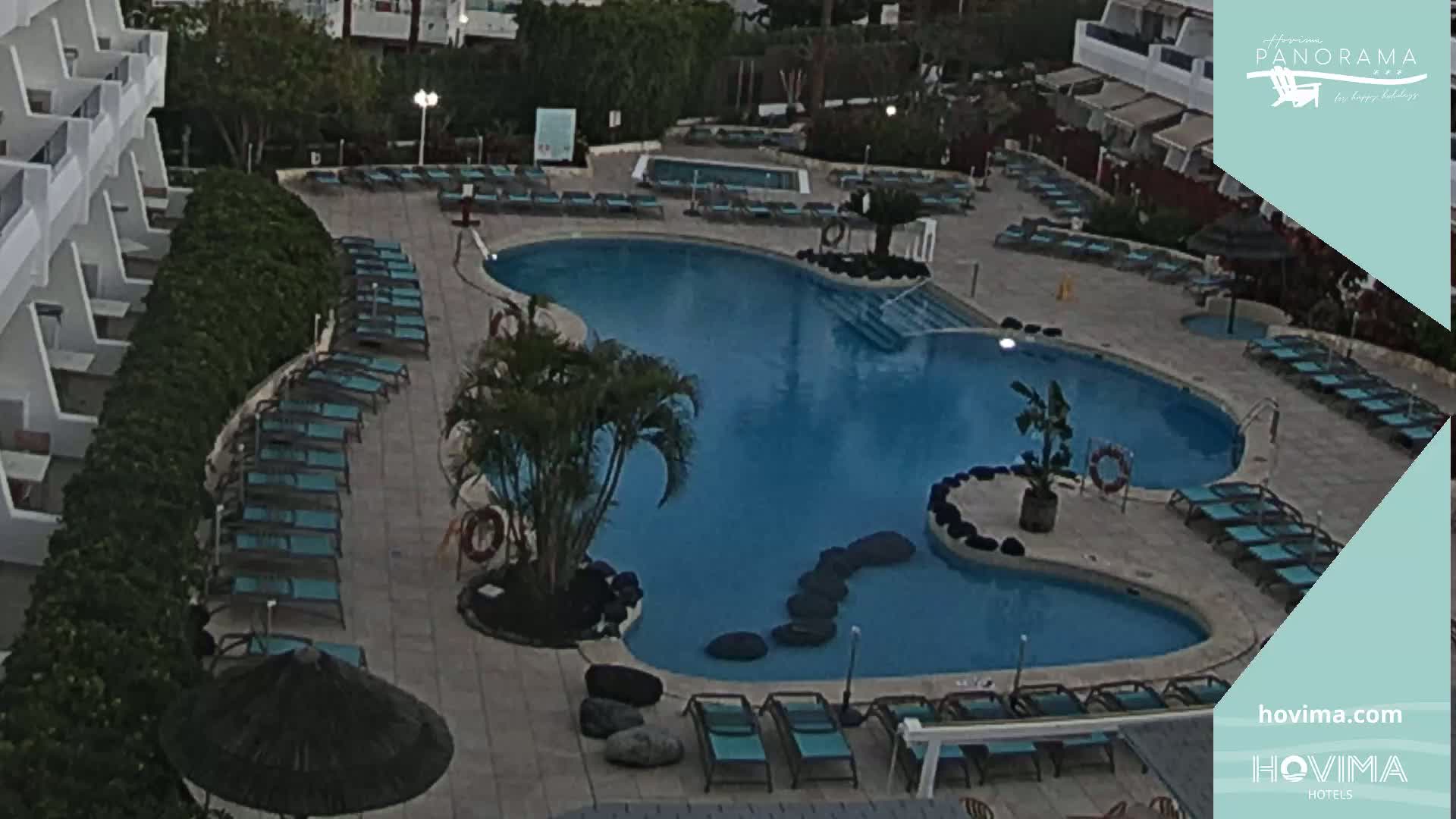 An aerial view shows a kidney-shaped swimming pool surrounded by lounge chairs next to a white building on a mostly cloudy day.