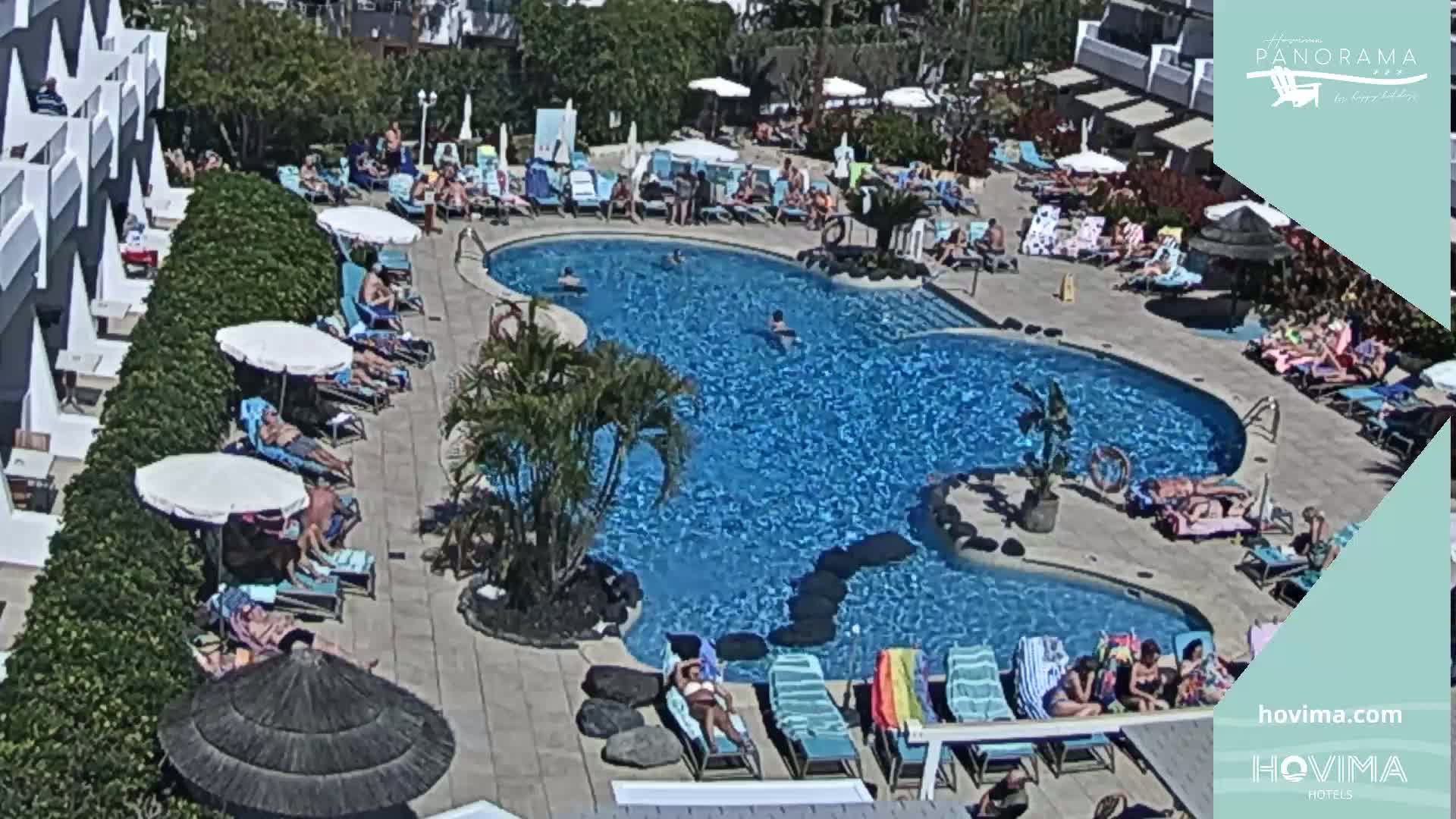 A sunny outdoor scene shows a resort pool with many people swimming and sunbathing around it, surrounded by lounge chairs and umbrellas.