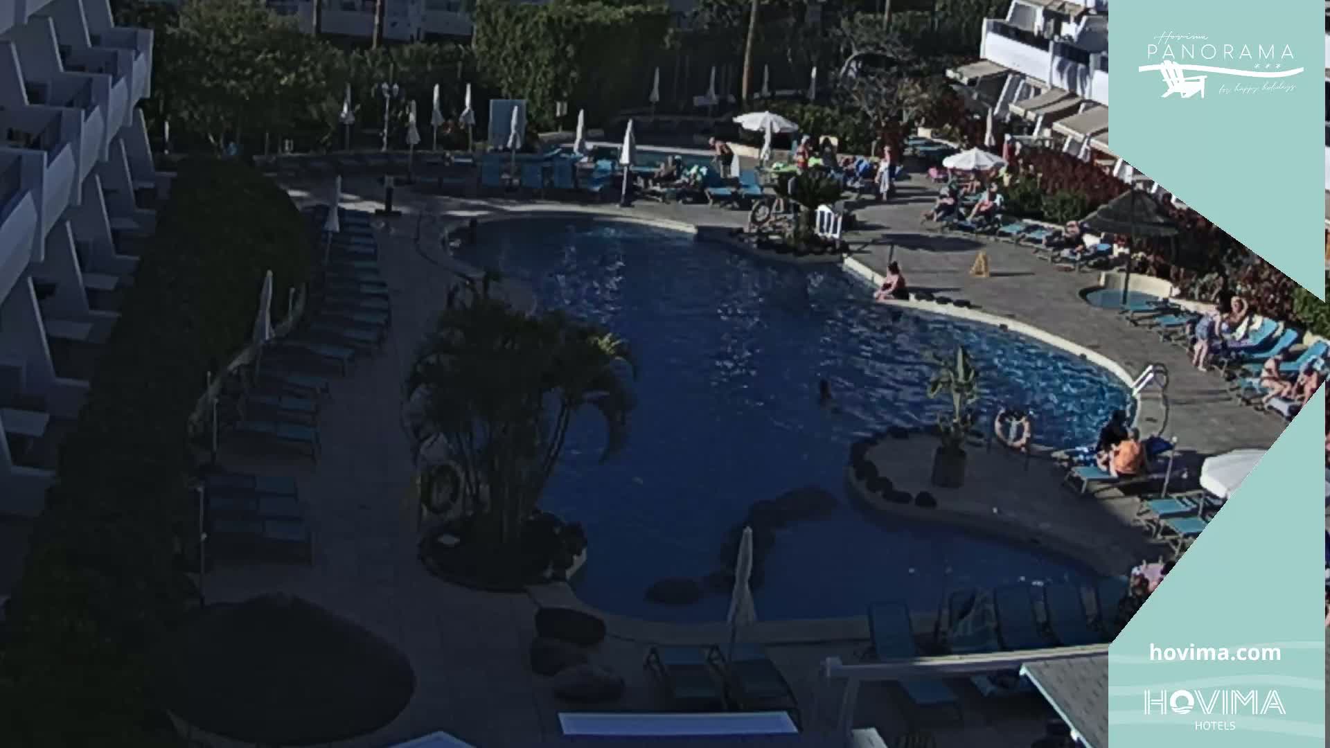 An outdoor swimming pool surrounded by lounge chairs and umbrellas, with people relaxing and swimming on a sunny day.