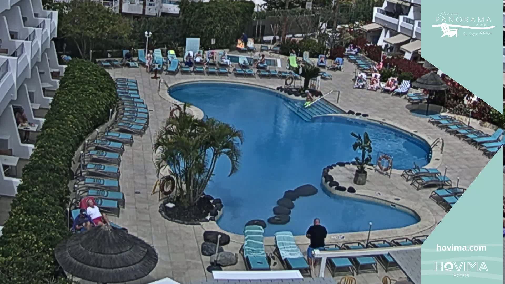 A resort pool area with numerous lounge chairs surrounding a kidney-shaped pool under sunny skies.
