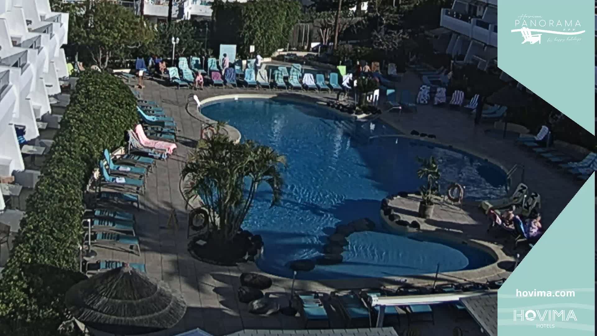 An outdoor swimming pool surrounded by lounge chairs and umbrellas, with several people relaxing around the pool on a sunny day.