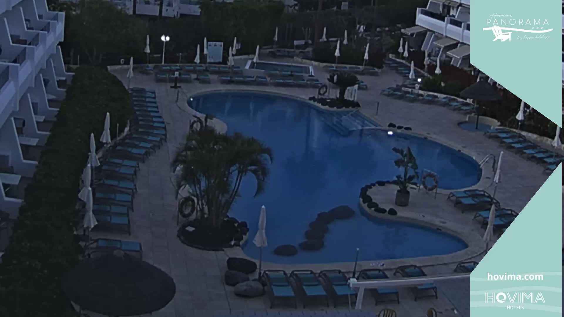 An outdoor swimming pool at night, surrounded by lounge chairs and umbrellas, next to a building.