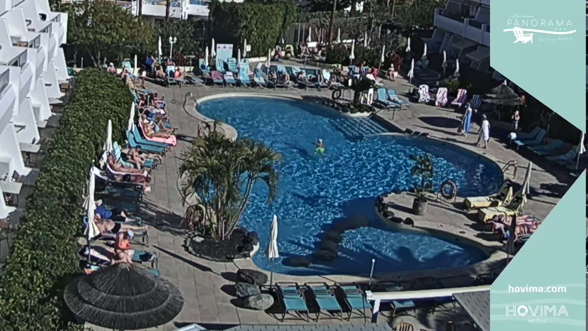A sunny outdoor pool area with many people lounging on chairs and in the water, surrounded by buildings and lush greenery.