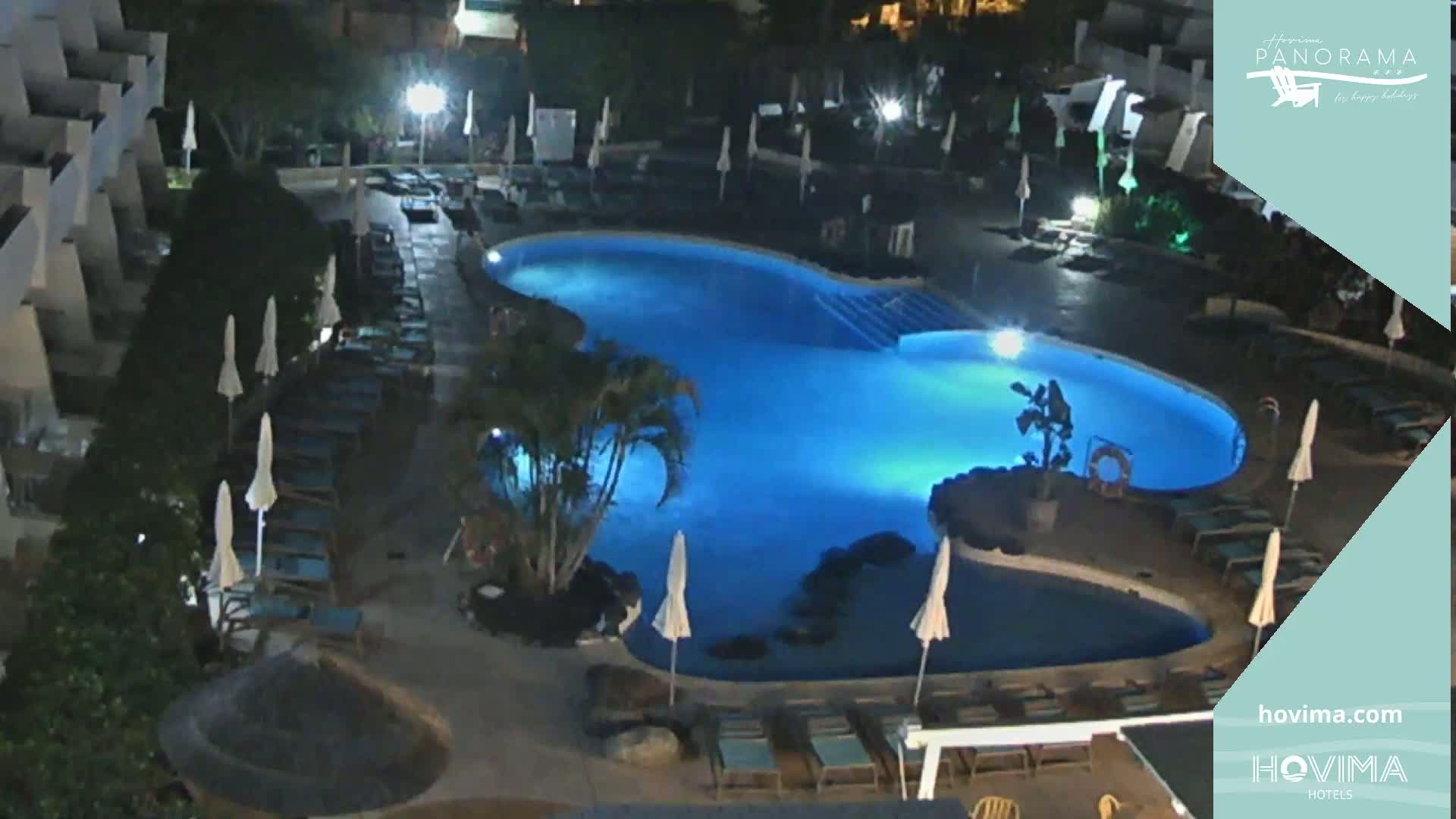 A brightly lit, kidney-shaped swimming pool surrounded by lounge chairs and umbrellas at night.